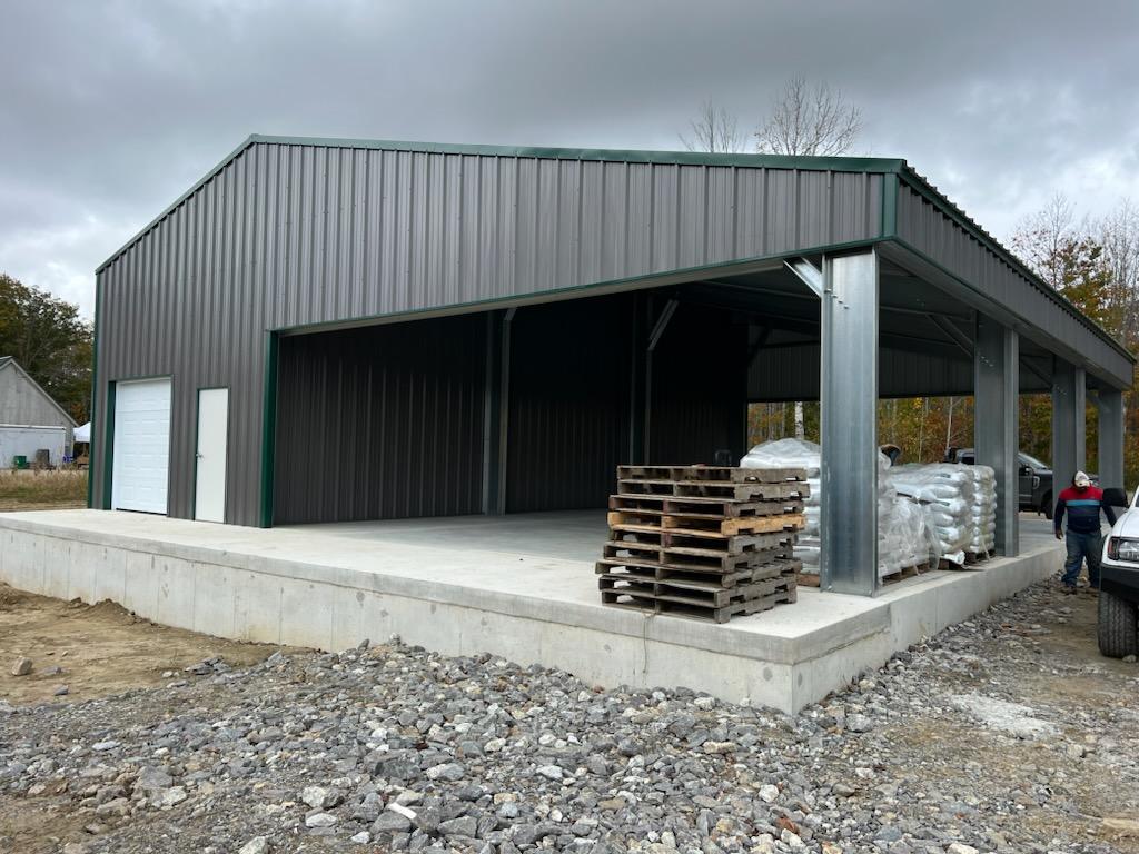 Metal building with a large open side, a closed section with a door and roll-up door, pallets stacked on a concrete foundation, under a cloudy sky.