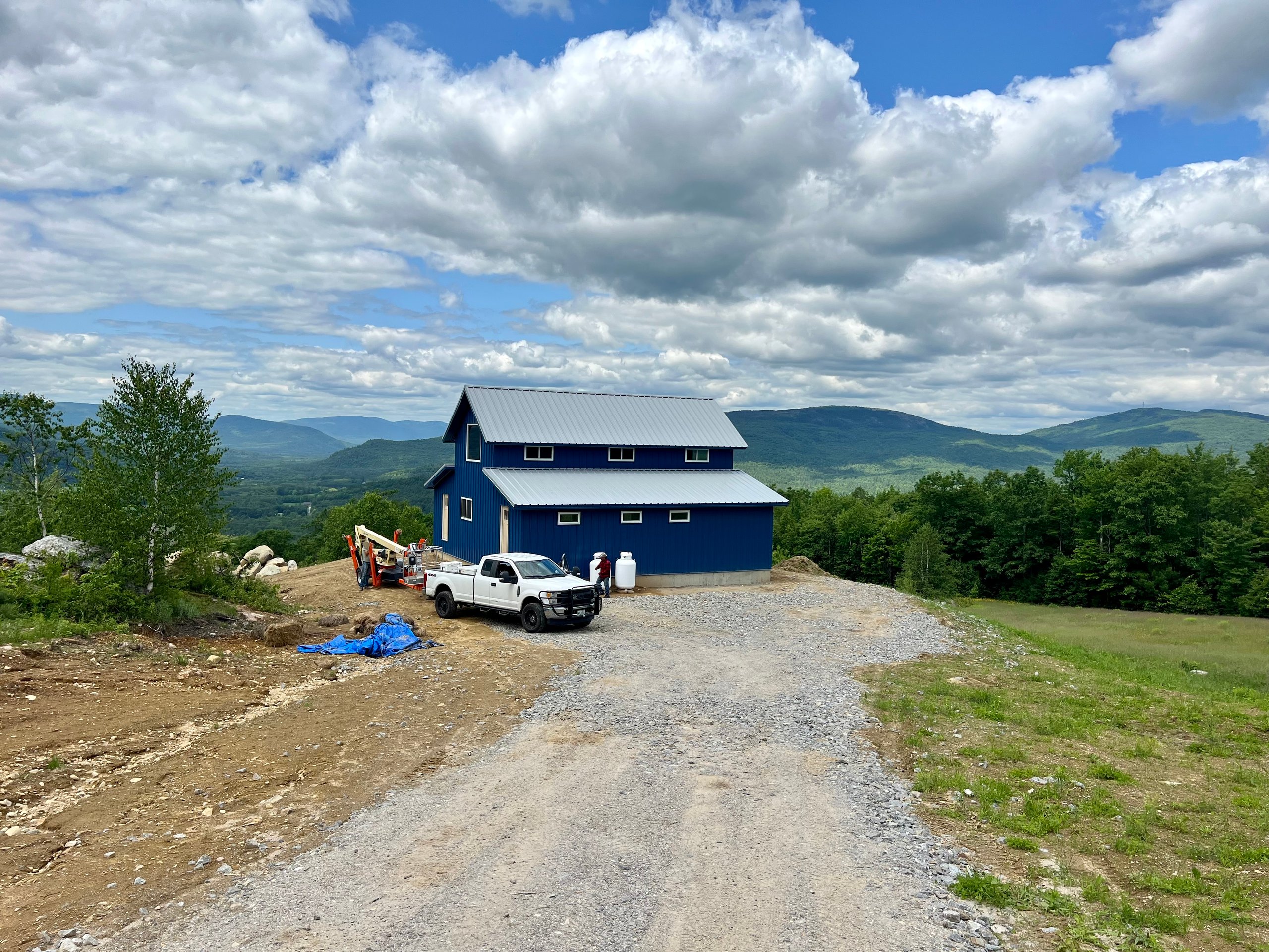 Blue two-story house with a metal roof on a gravel driveway with a white pickup truck and workers, surrounded by green trees and mountains under a cloudy sky.