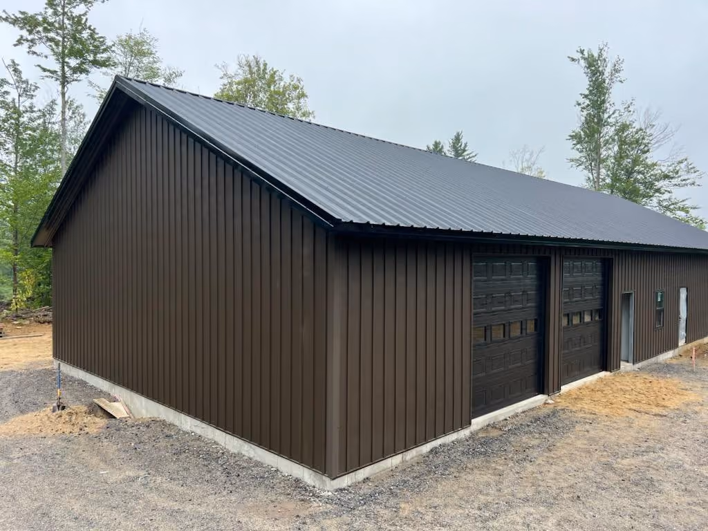 Large brown metal building with two garage doors and a small side door on a gravel and dirt lot surrounded by sparse trees.
