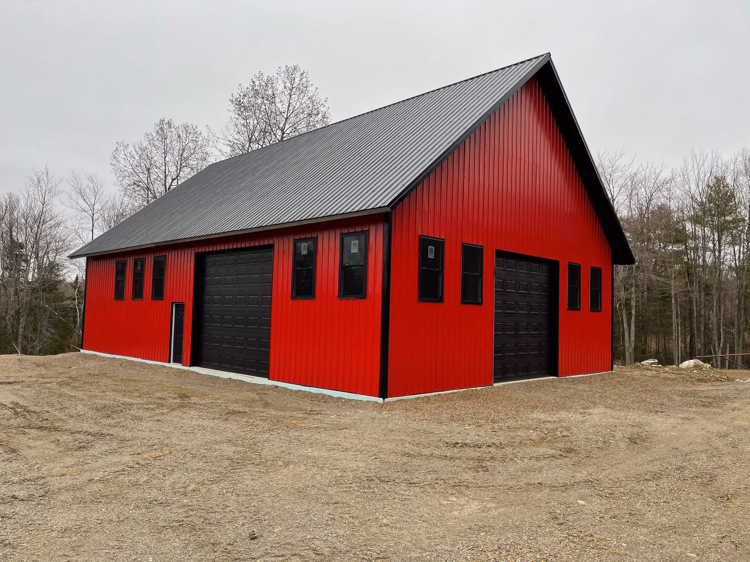 Large red metal barn with black garage doors and multiple windows on a dirt clearing surrounded by leafless trees.