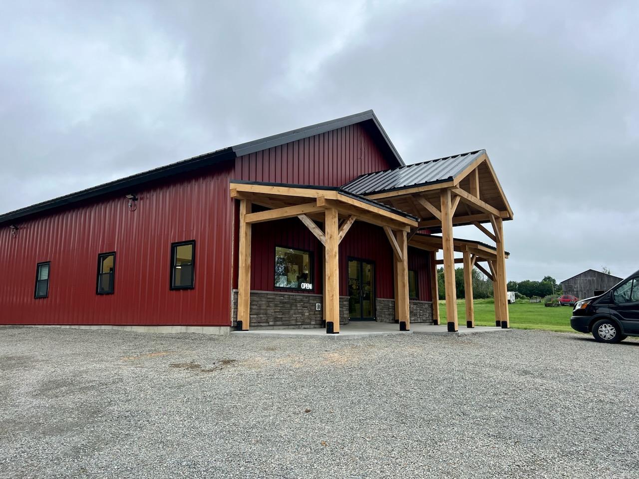 Red metal building with wooden porch structure and an open sign on the window, gravel parking in front under cloudy sky.