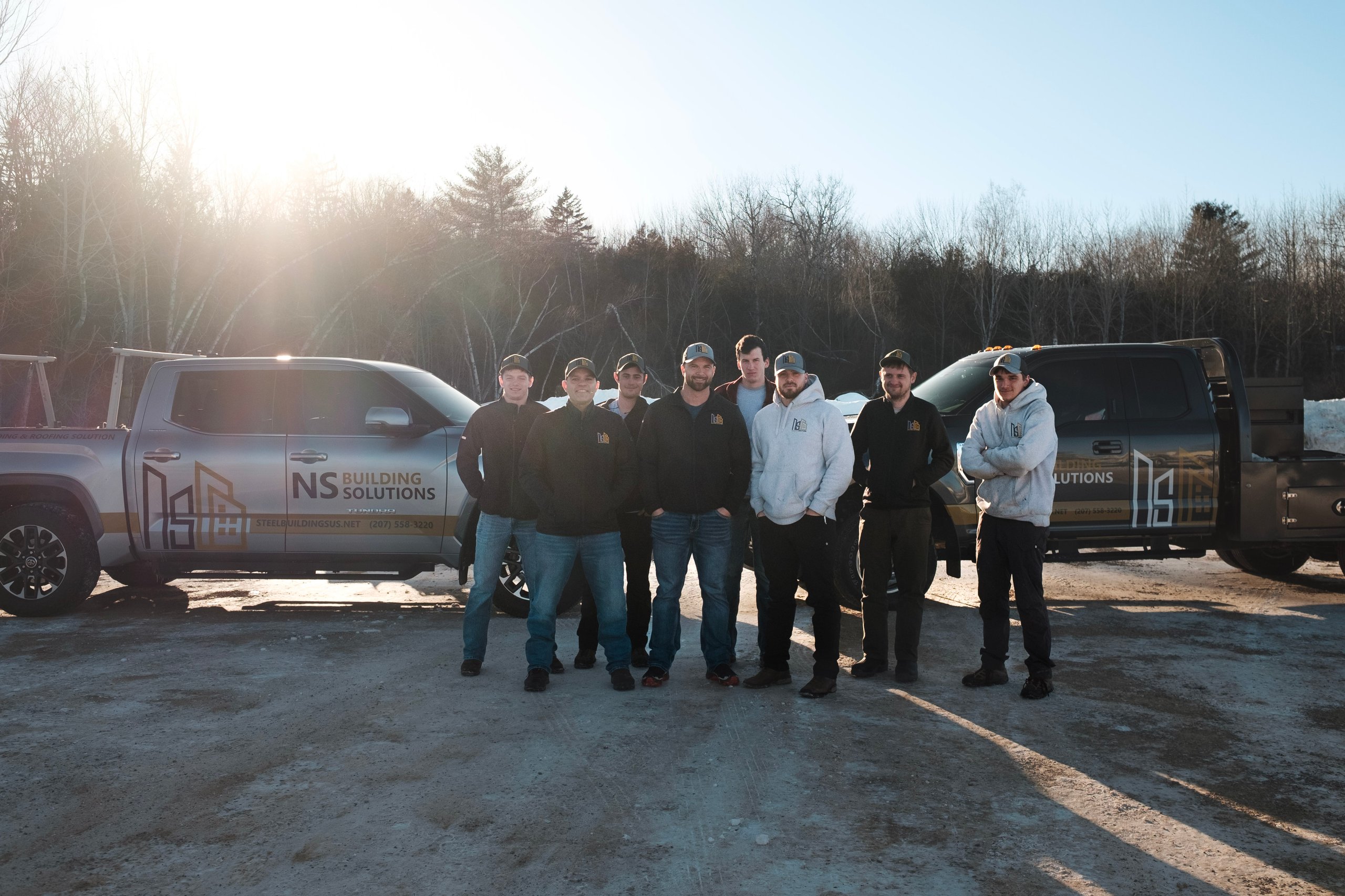 Eight men standing outdoors in front of two NS Building Solutions trucks with trees and bright sunlight in the background.
