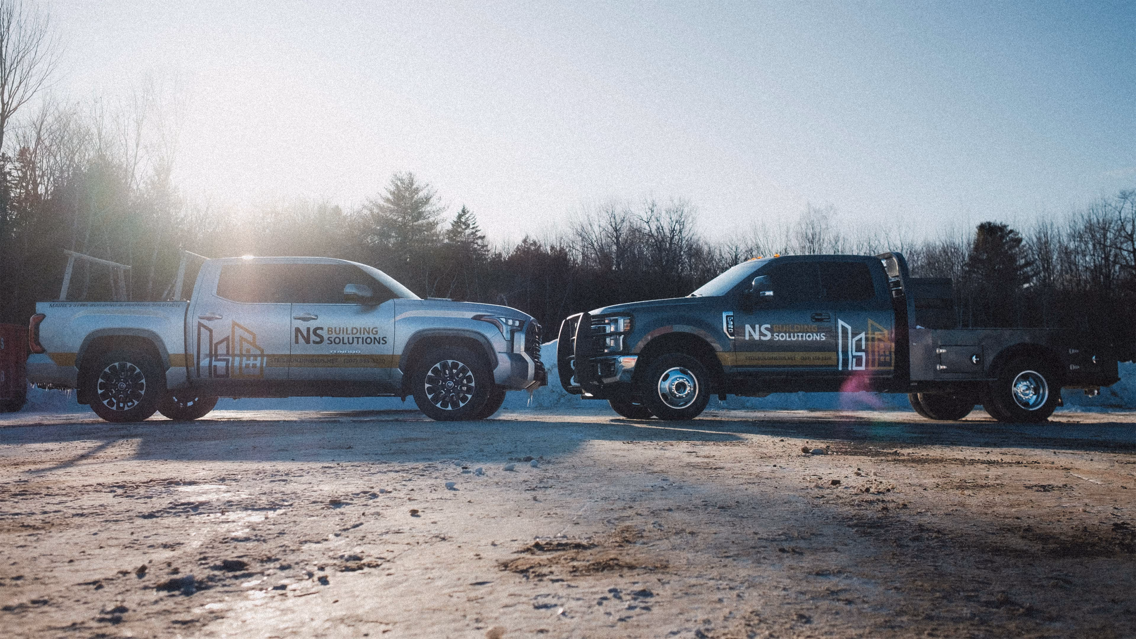 Two NS Building Solutions pickup trucks parked facing each other on a snowy ground with a background of leafless trees and a bright sun.