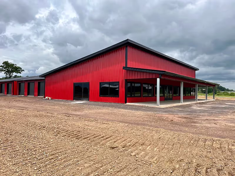 Large red metal building with single sloped roof and covered porch on a gravel lot under a cloudy sky.
