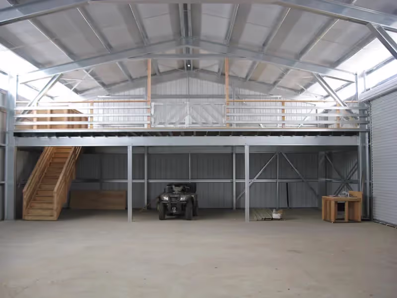 Interior of a large metal warehouse with a wooden staircase leading to a mezzanine and an ATV parked underneath.