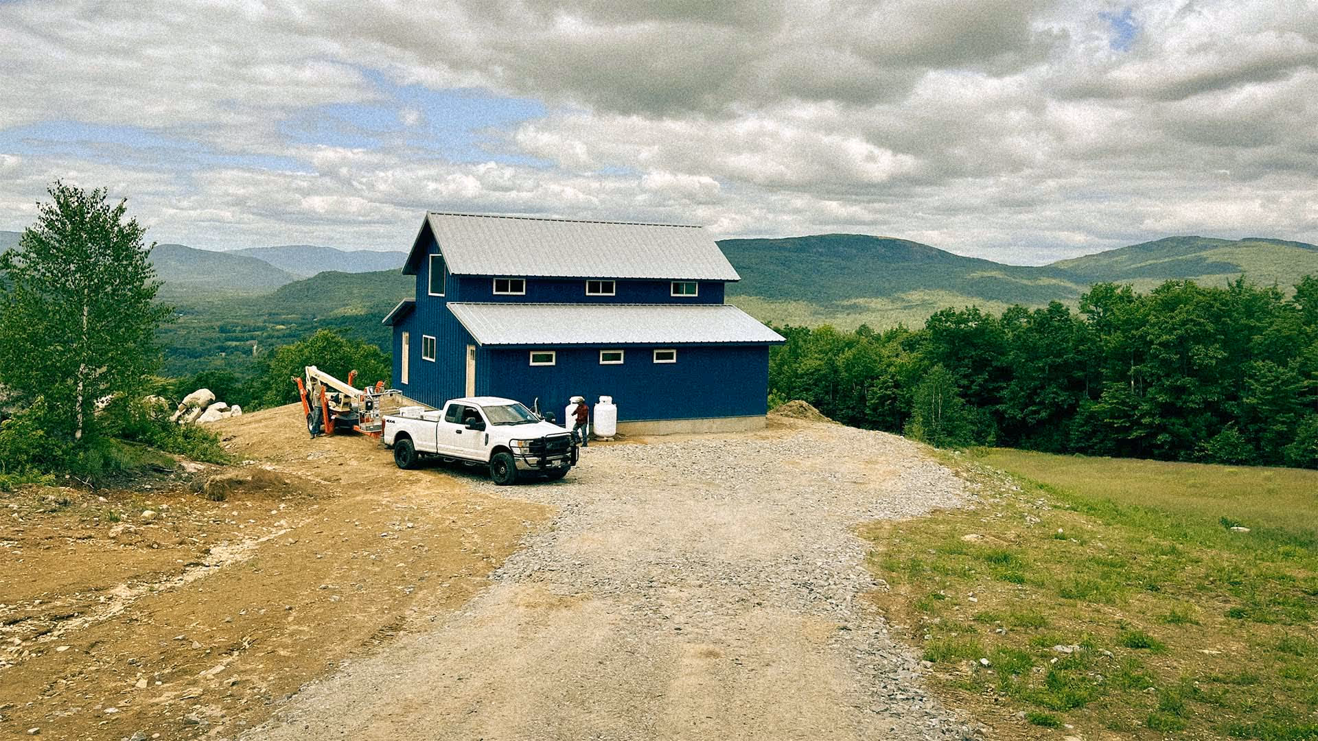 Blue barn-style house with metal roof on a rural gravel lot surrounded by green trees and mountains under a cloudy sky.