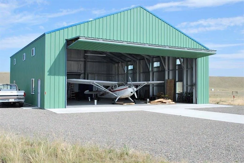 Small white airplane parked inside a green metal hangar with the door open, next to a vintage white pickup truck.