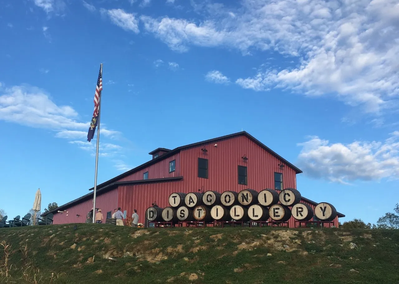 Red barn-style building on a grassy hill with large barrels spelling out 'Taconic Distillery' and an American flag on a pole.