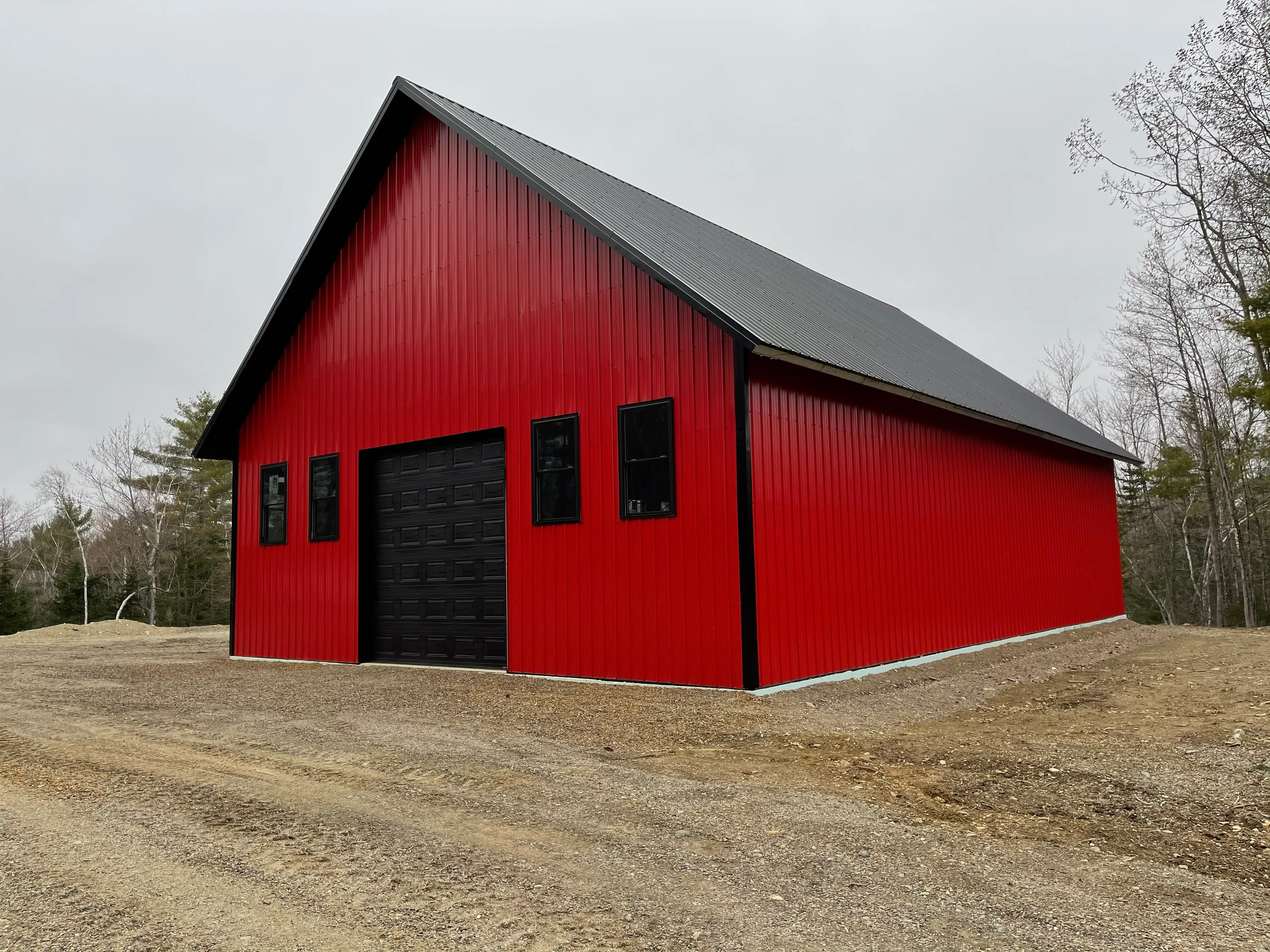 Large red metal building with black roof, black garage door, and four black-framed windows, surrounded by dirt and trees.