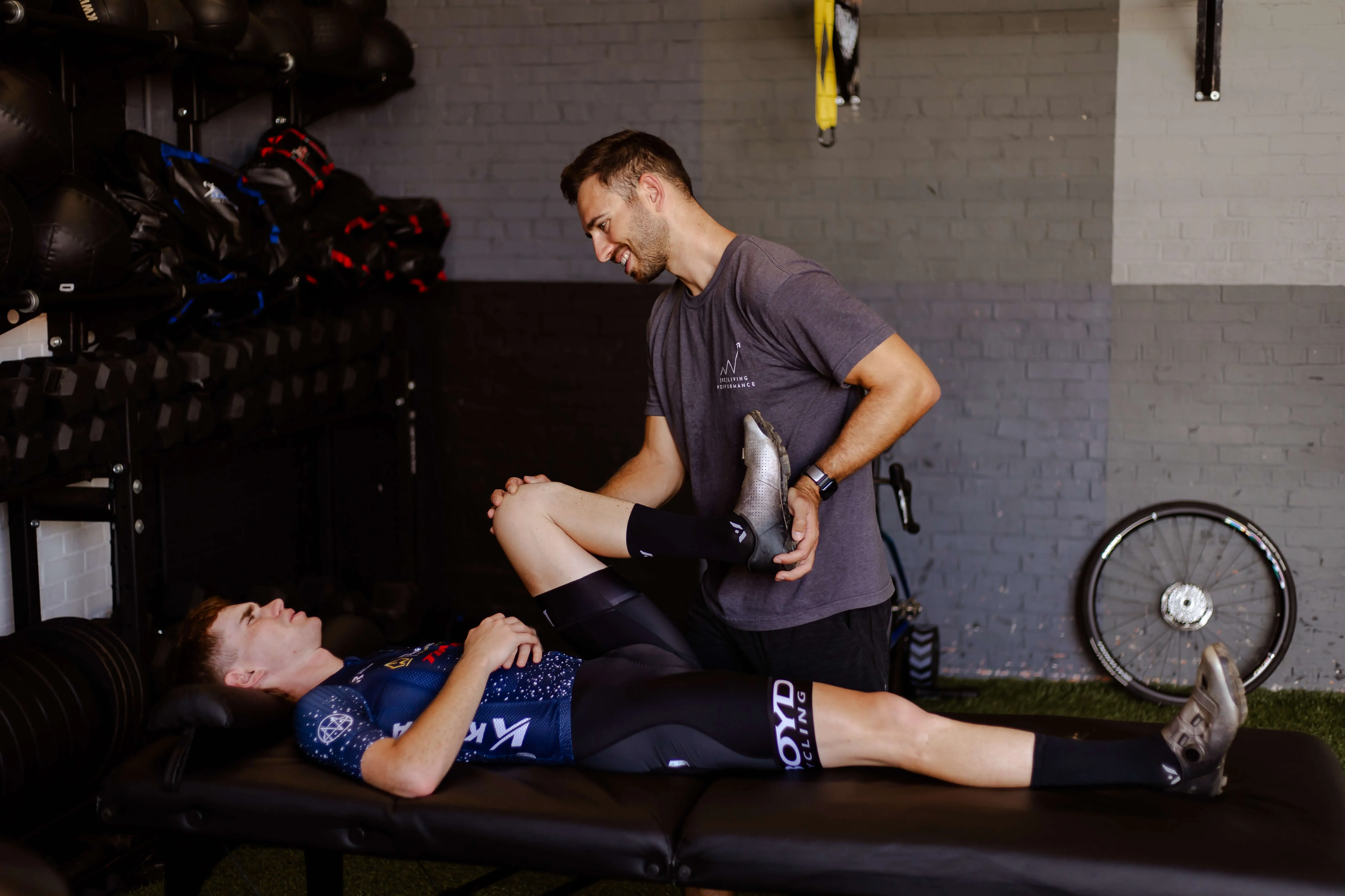 A man in a gray shirt assists a cyclist lying on a massage table by stretching his leg in a gym setting.