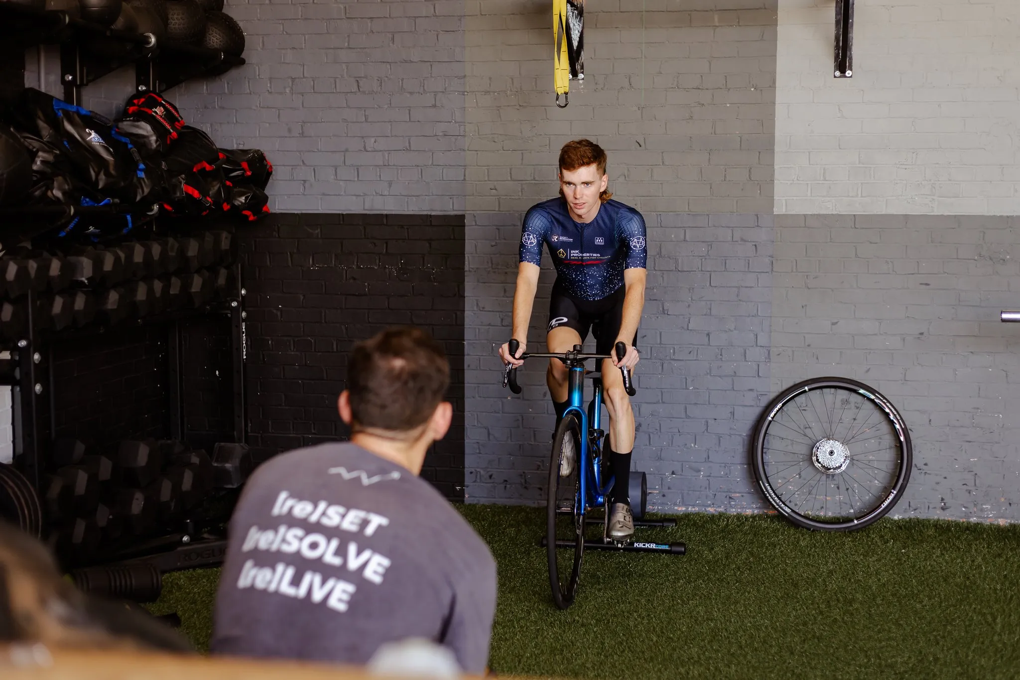 Cyclist wearing a blue jersey training on a stationary bike trainer indoors while another person watches.
