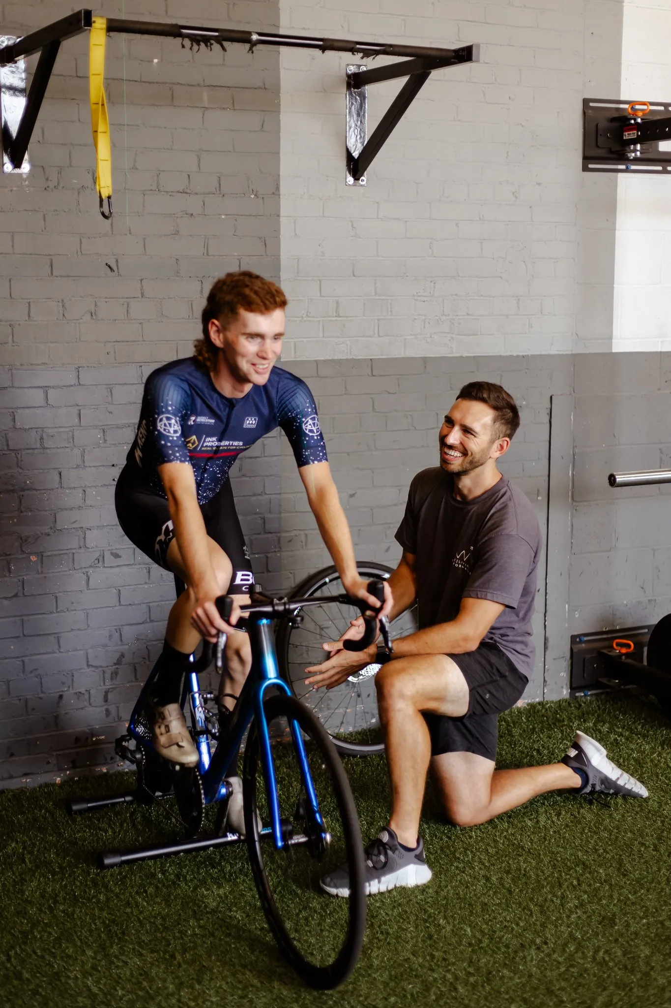 Cyclist riding a stationary bike indoors while a coach kneels beside him, smiling and providing encouragement.