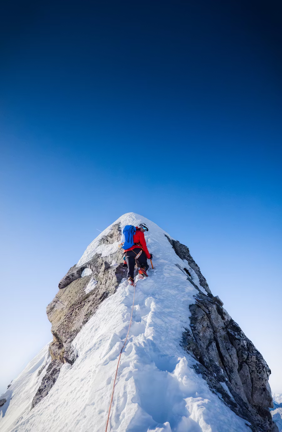 Climber standing with arms outstretched on a snowy summit, rope trailing behind, vast alpine panorama below