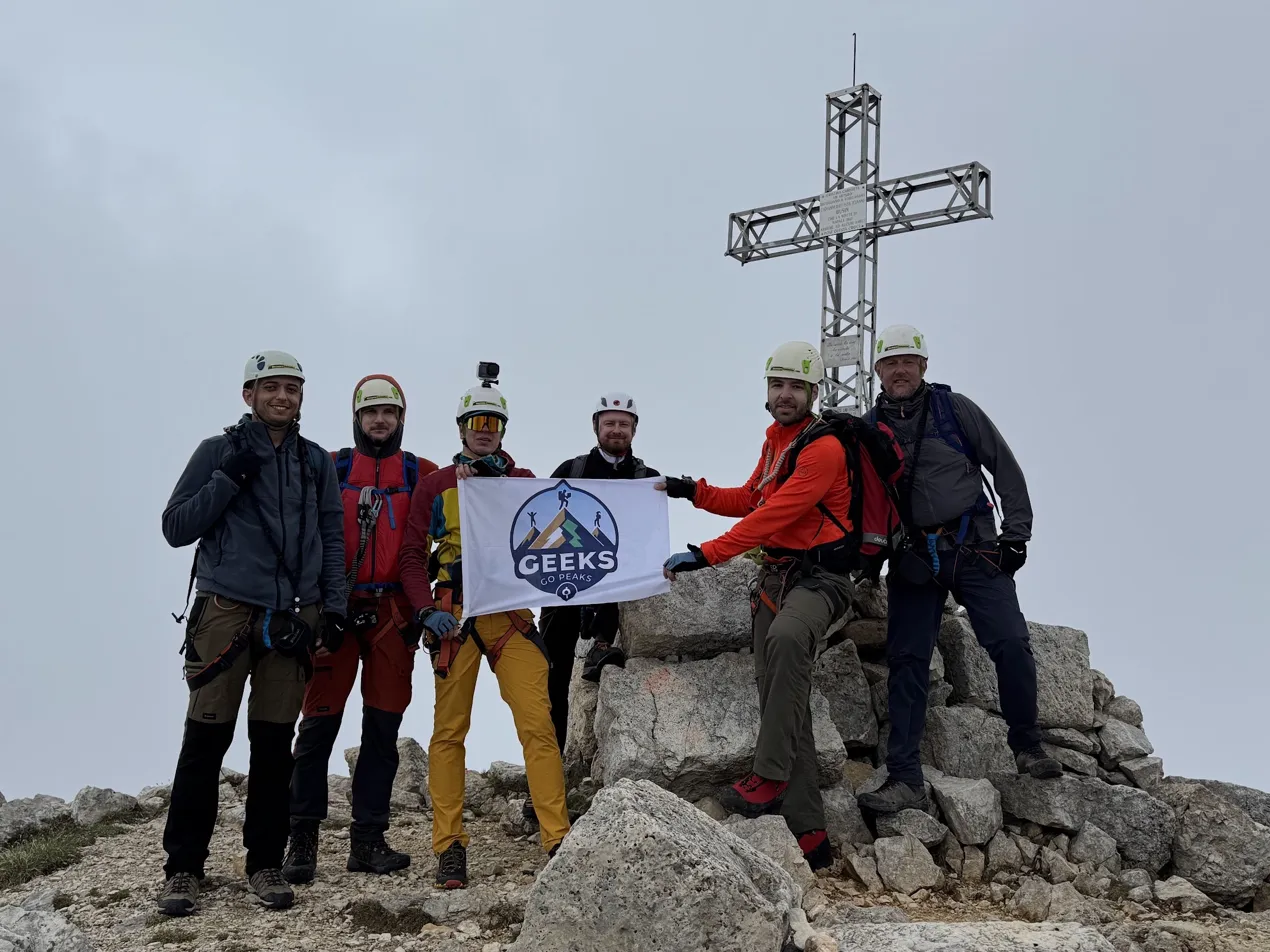 Geeks Go Peaks group holding the GGP flag at the summit cross — six climbers in helmets after completing the ridge