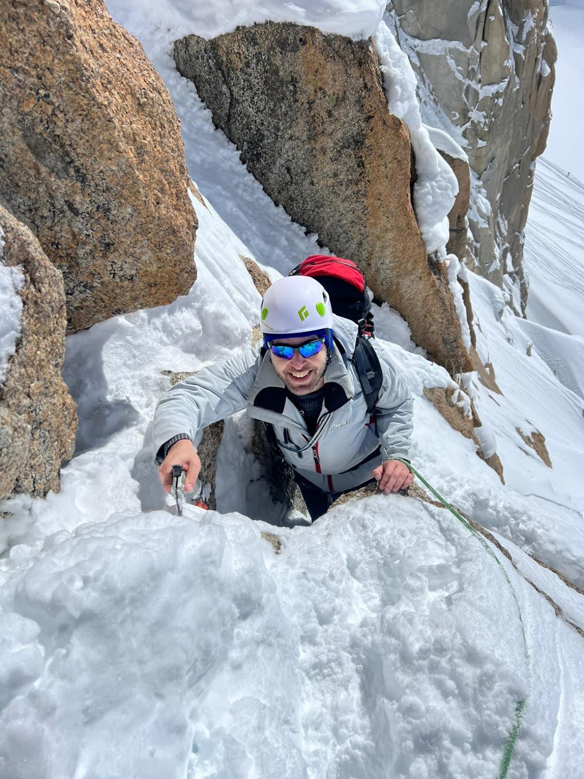 Climber ascending a mixed rock-and-snow gully on the Arête des Cosmiques, smiling up at the camera