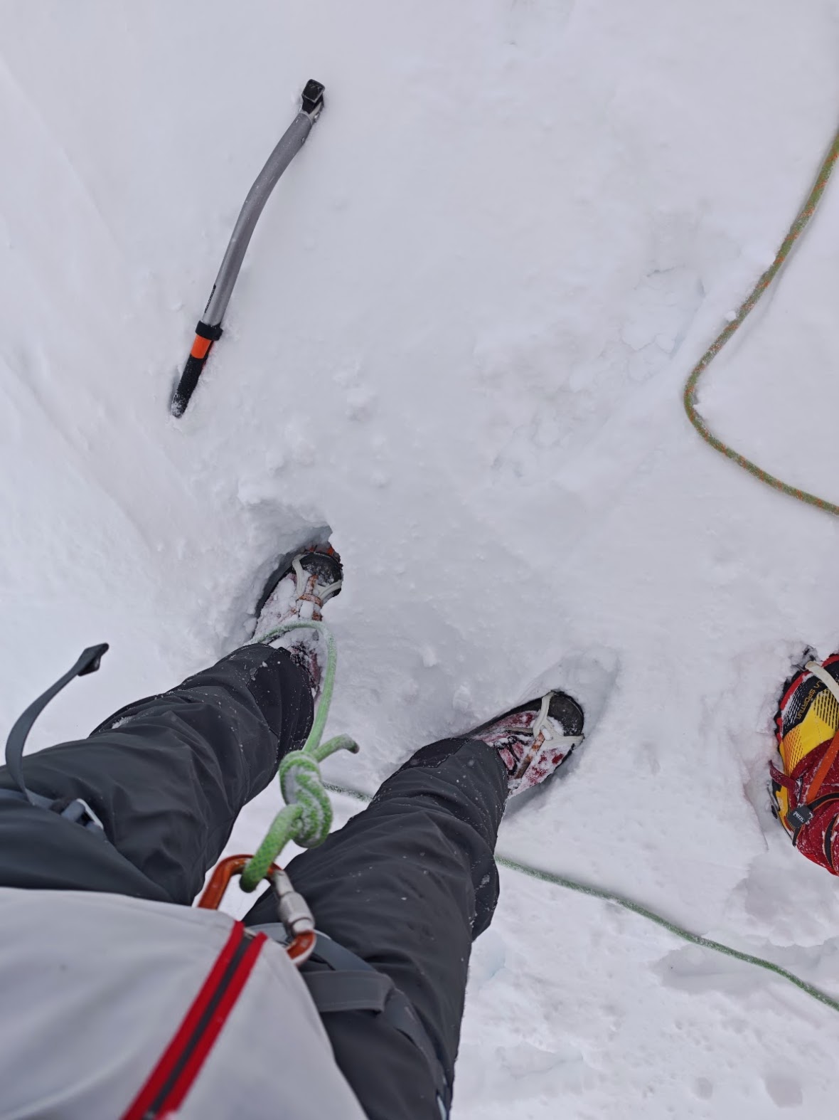 Team member wading through fresh deep snow in whiteout conditions, rope visible