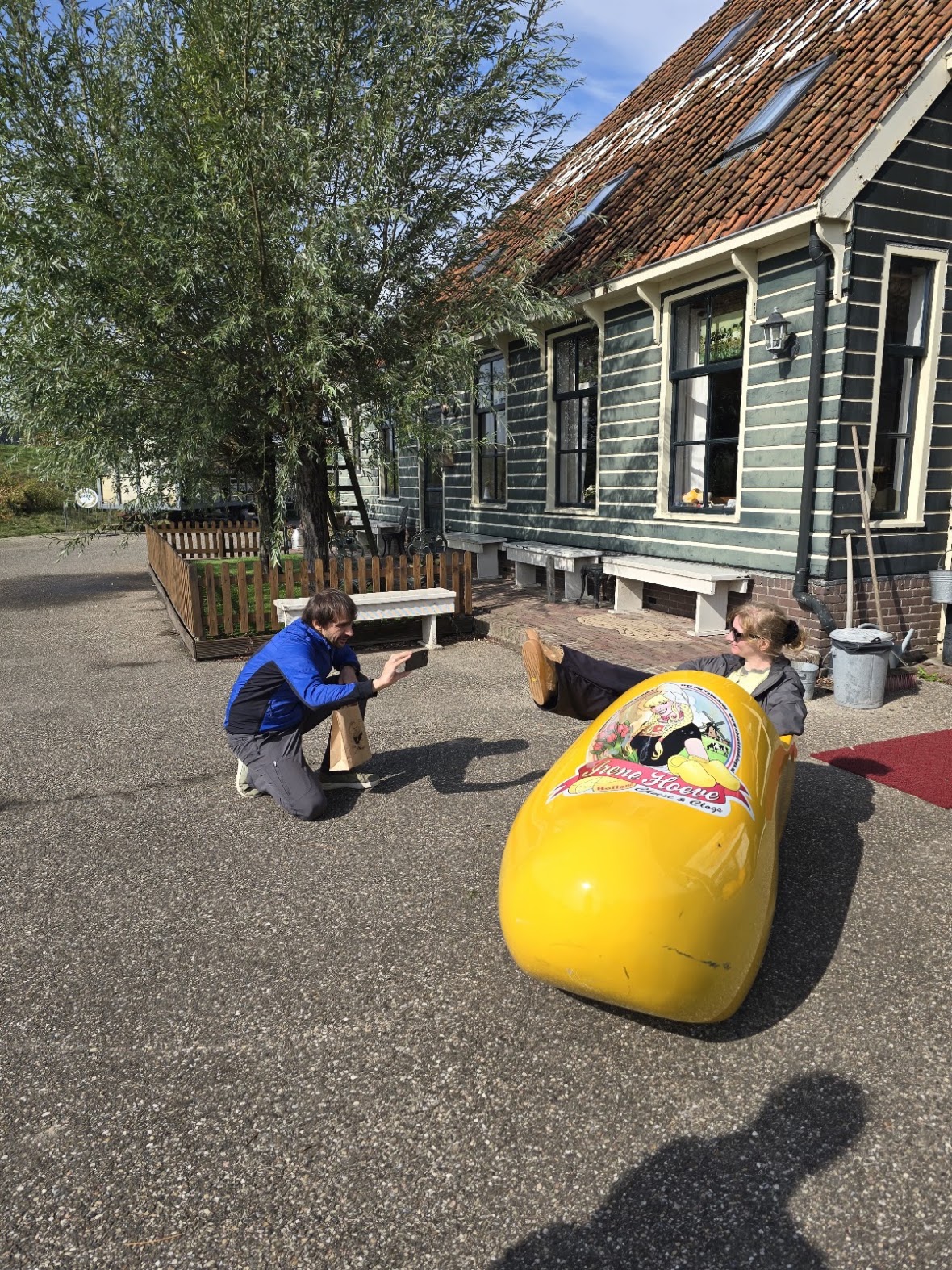 Two participants posing with an oversized wooden clog outside a farmhouse