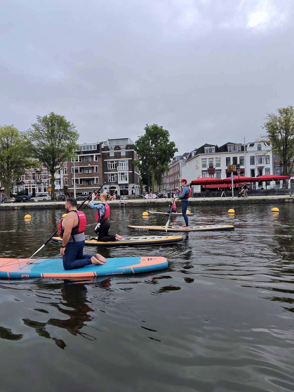 Two paddlers kneeling on stand-up paddleboards on the Amstel with Amsterdam buildings behind them