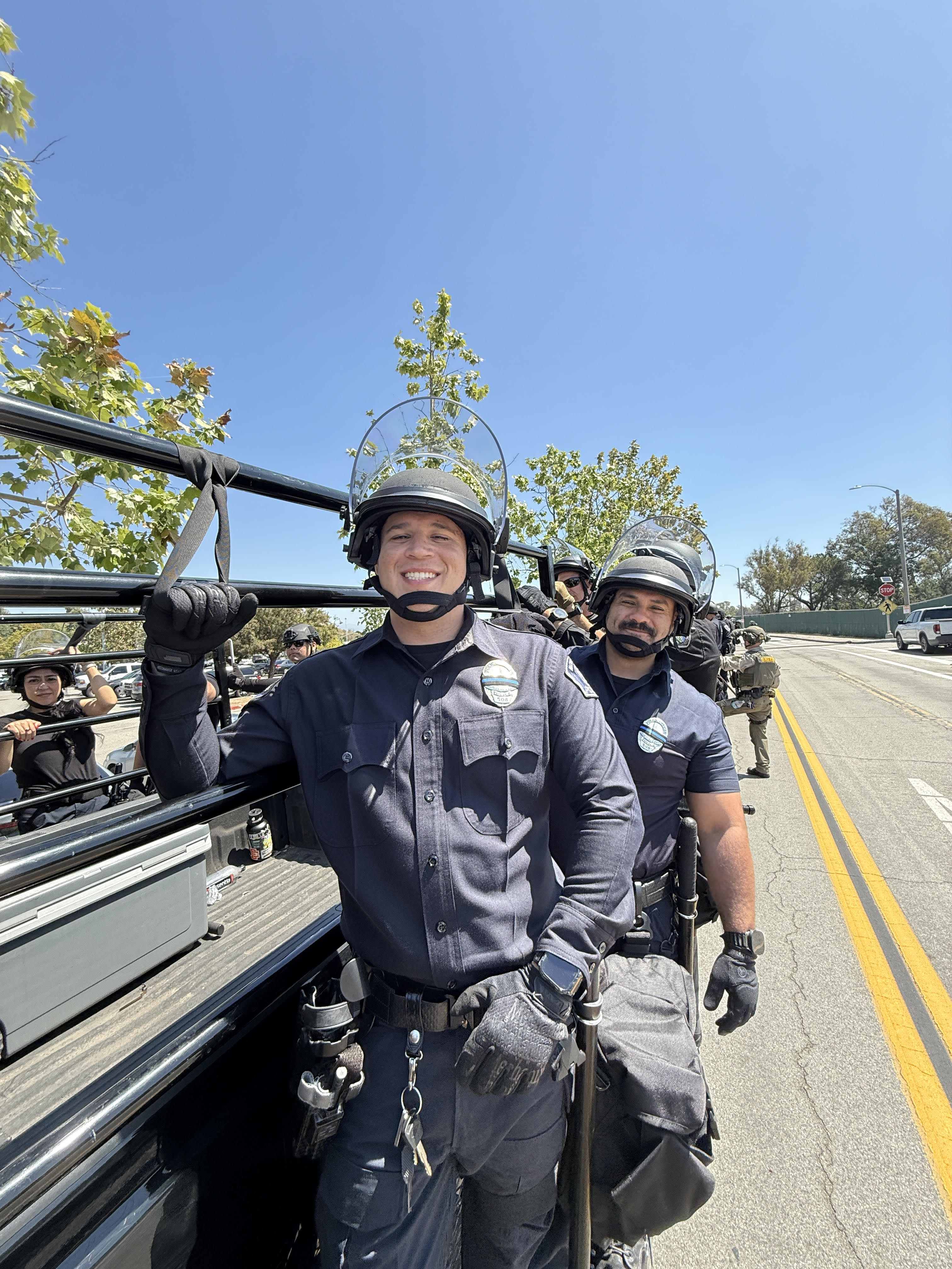 Smiling police officers in protective gear standing in the back of a vehicle on a sunny street.