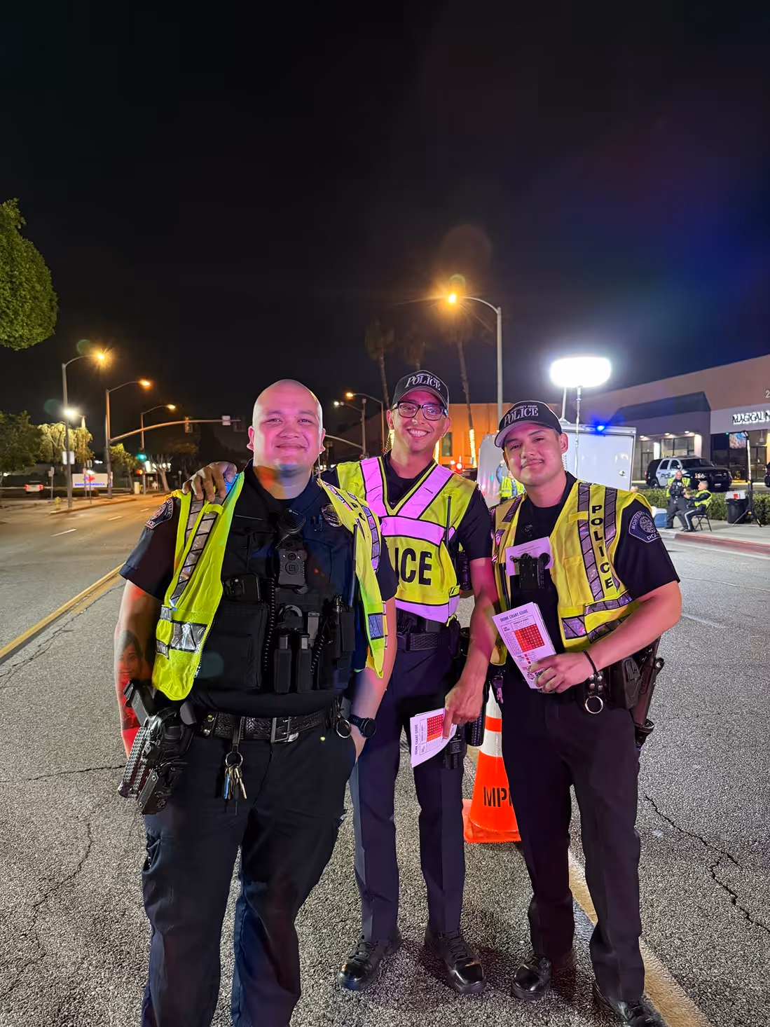 Three police officers in reflective vests posing on a street at night, smiling and holding papers.