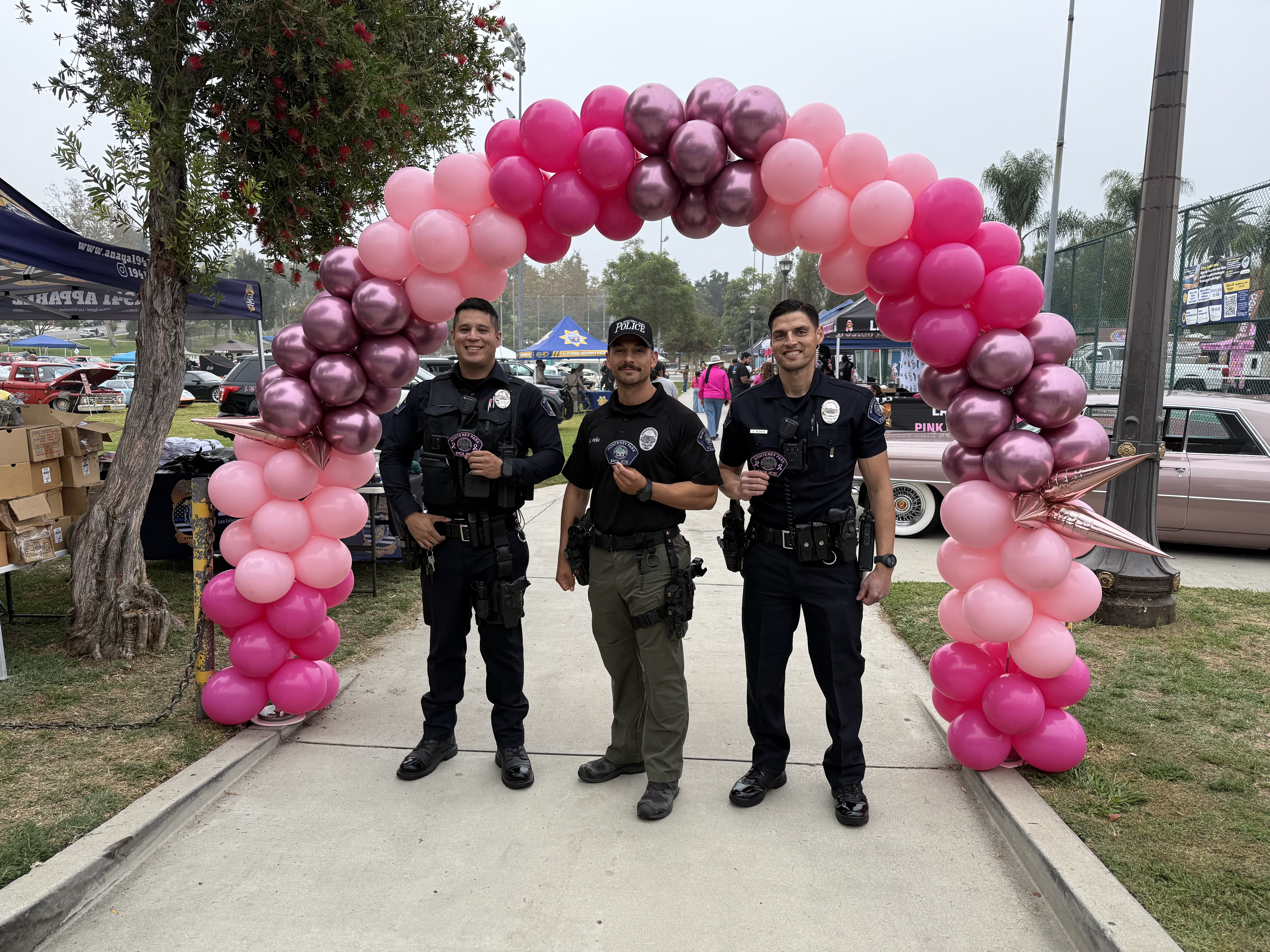 Three police officers standing under a pink and purple balloon arch at an outdoor event.