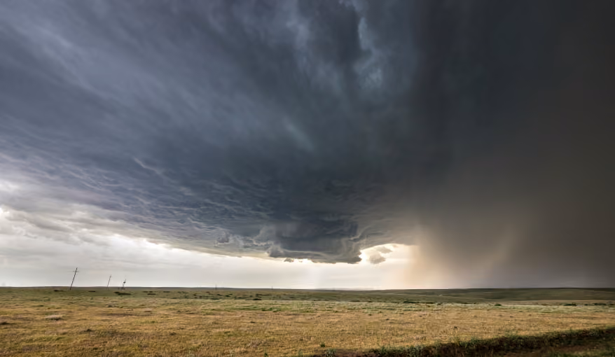 Dark storm clouds gathering over a flat grassy plain with rain falling in the distance.