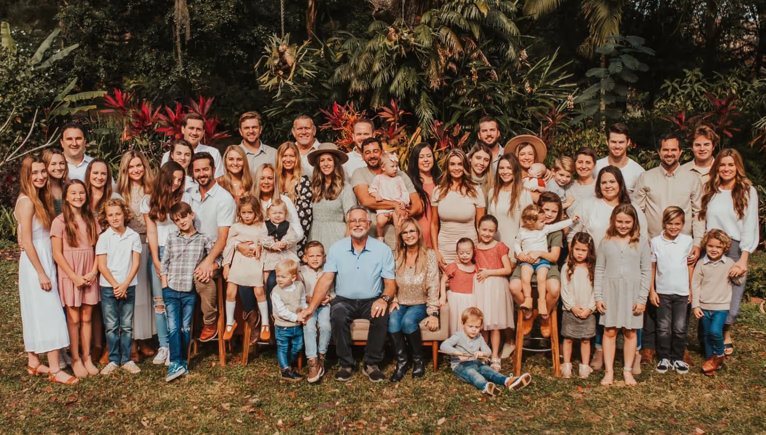 Large multi-generation family posing outdoors on grass with tropical plants and trees in the background.