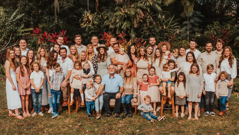 Large multi-generation family posing outdoors on grass with tropical plants and trees in the background.