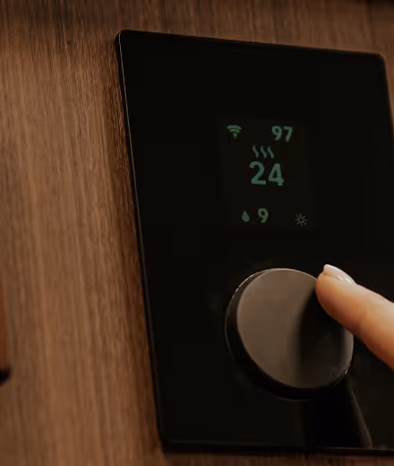 Close-up of a hand adjusting a black digital thermostat with temperature set to 24 degrees on a wooden wall.