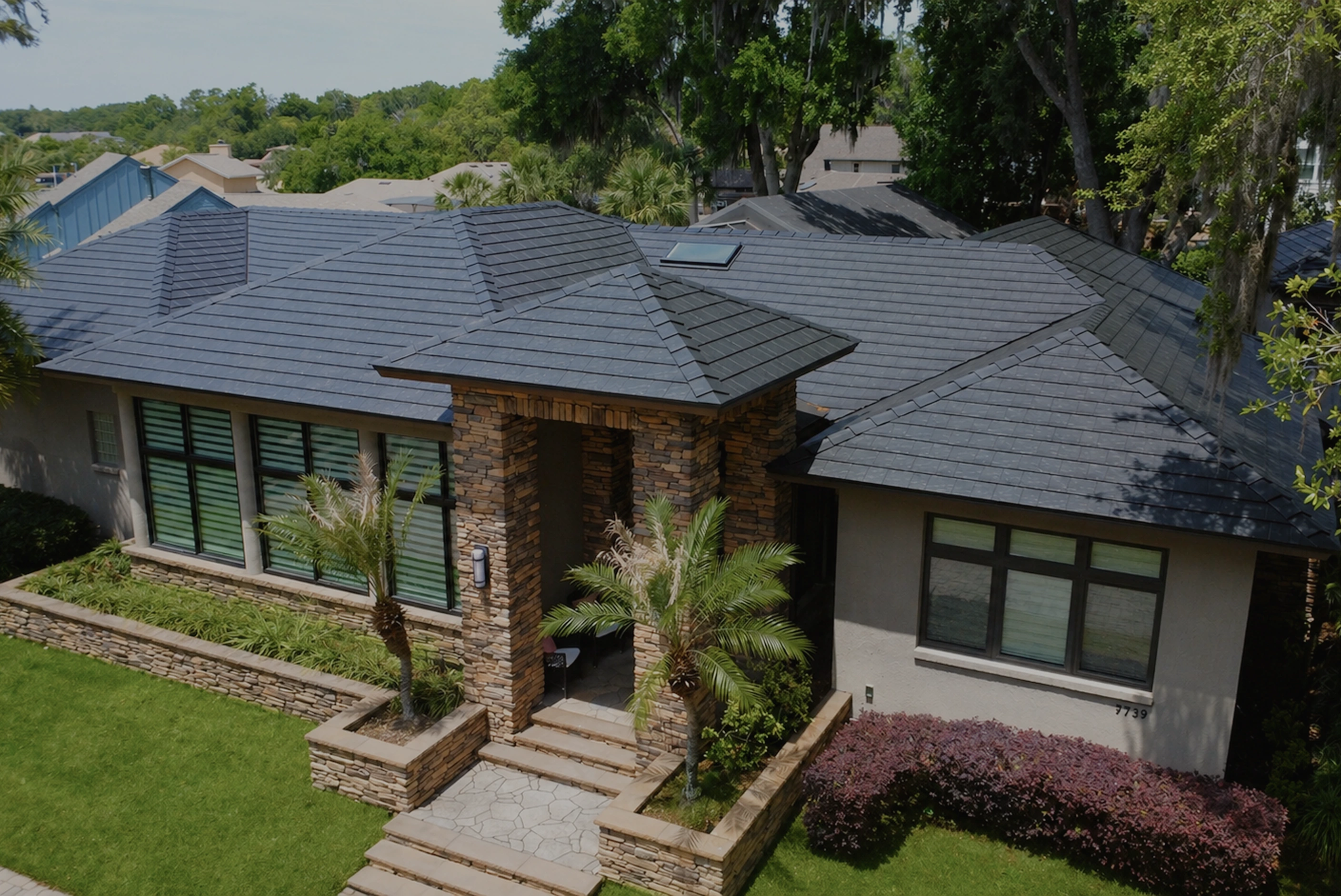 Modern single-story house with dark tiled roof, stone columns, palm trees, and landscaped front yard.