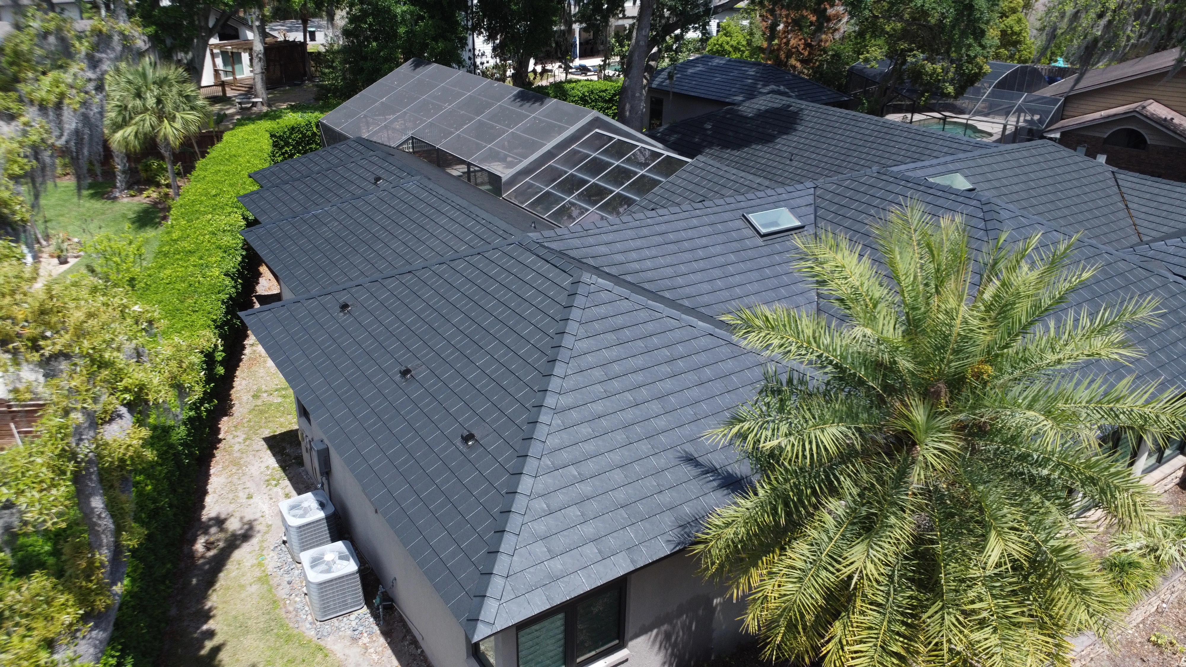Aerial view of a modern house with dark slate tile roofing surrounded by greenery and palm trees.