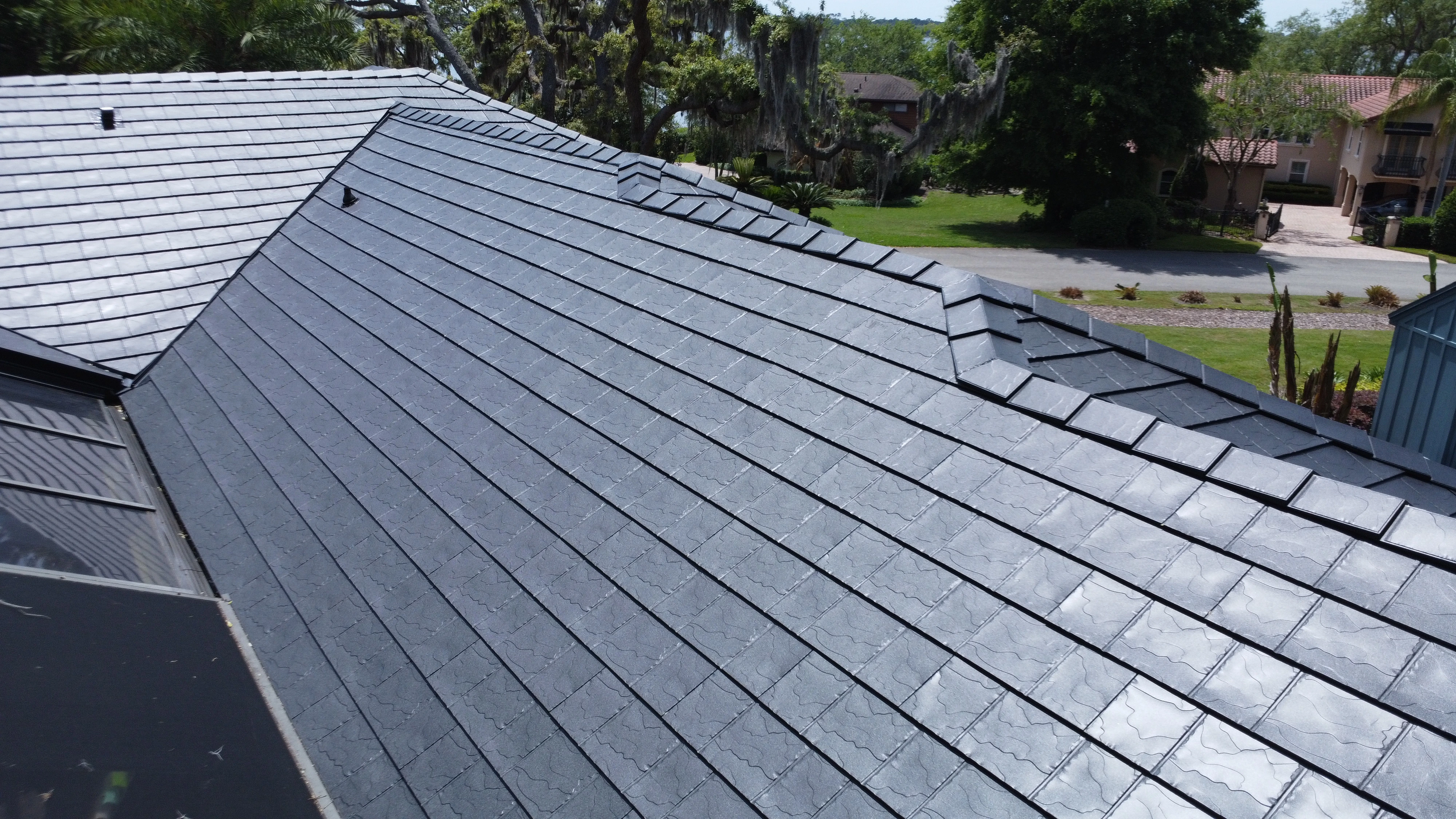 Close-up view of a large residential roof with dark gray slate tiles, overlooking green lawns and neighboring houses.