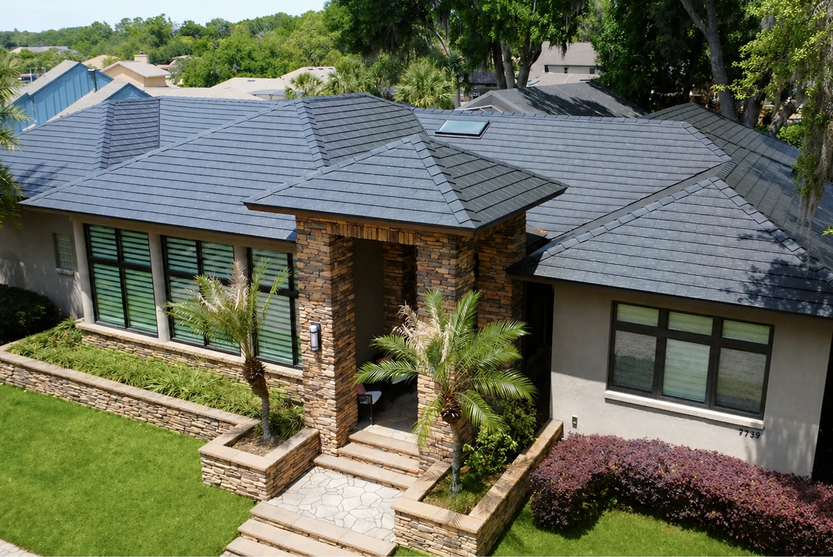 Modern single-story house with a dark tiled roof, stone facade entrance, large windows, and palm trees in the front yard.