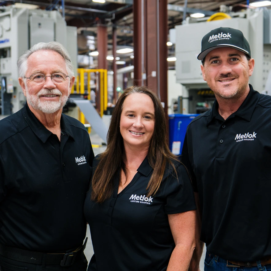 Three people in black Metlok Lifetime Roofing shirts smiling inside an industrial facility.
