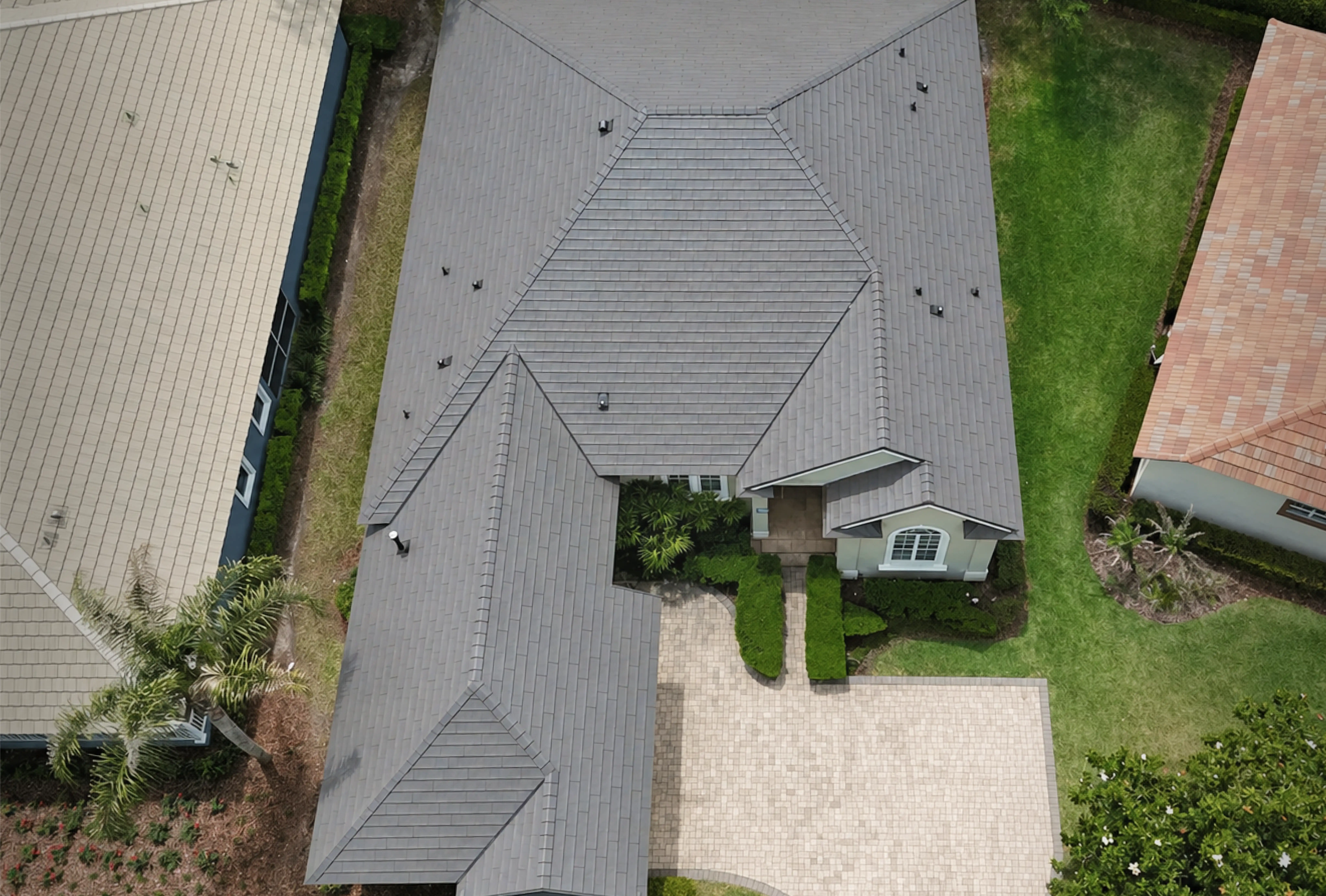 Aerial view of a residential house with a grey tiled roof, a driveway, green lawn, and neighboring houses.