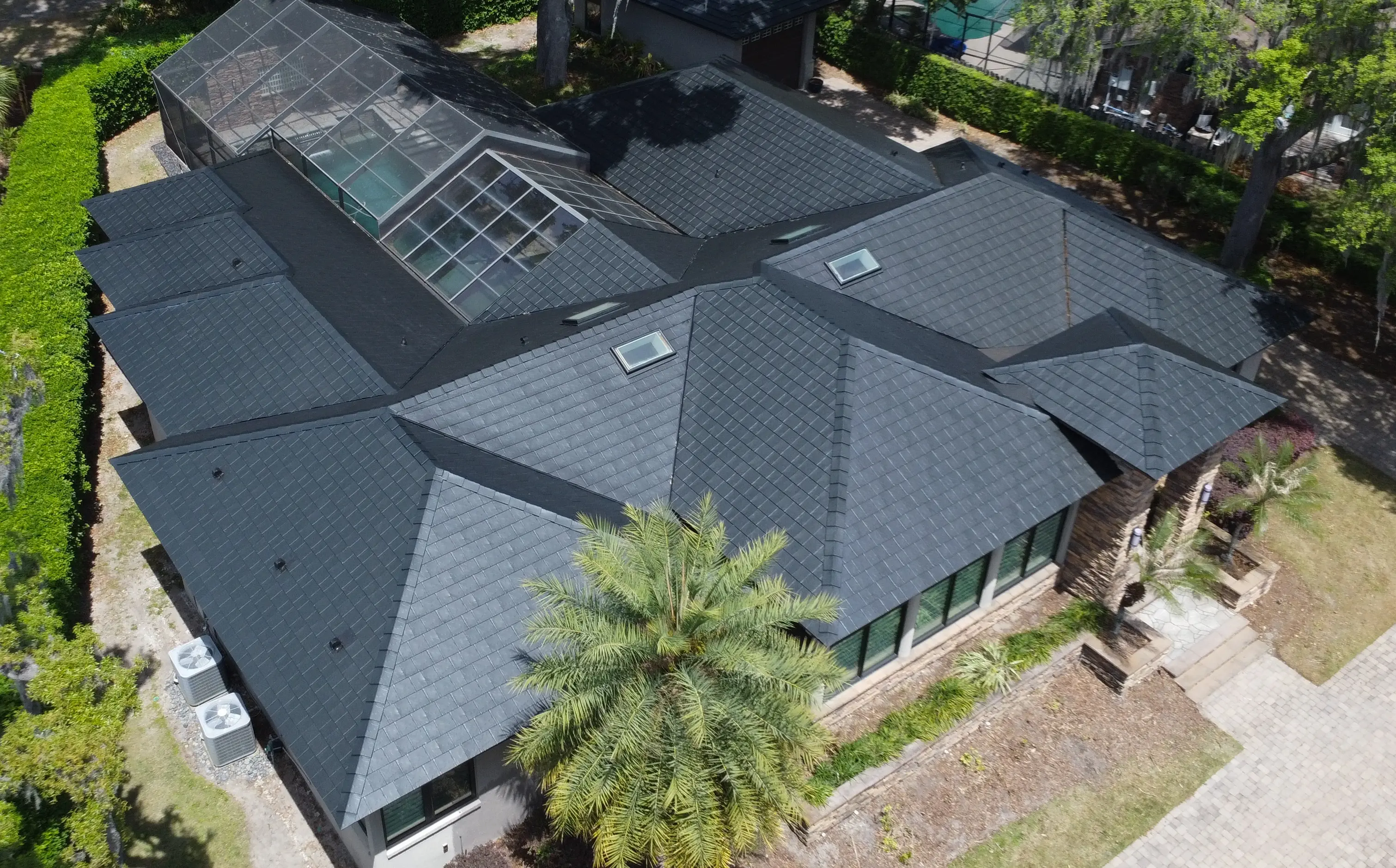 Aerial view of a large house with a black, multi-faceted slate roof, skylights, and a screened-in pool enclosure.