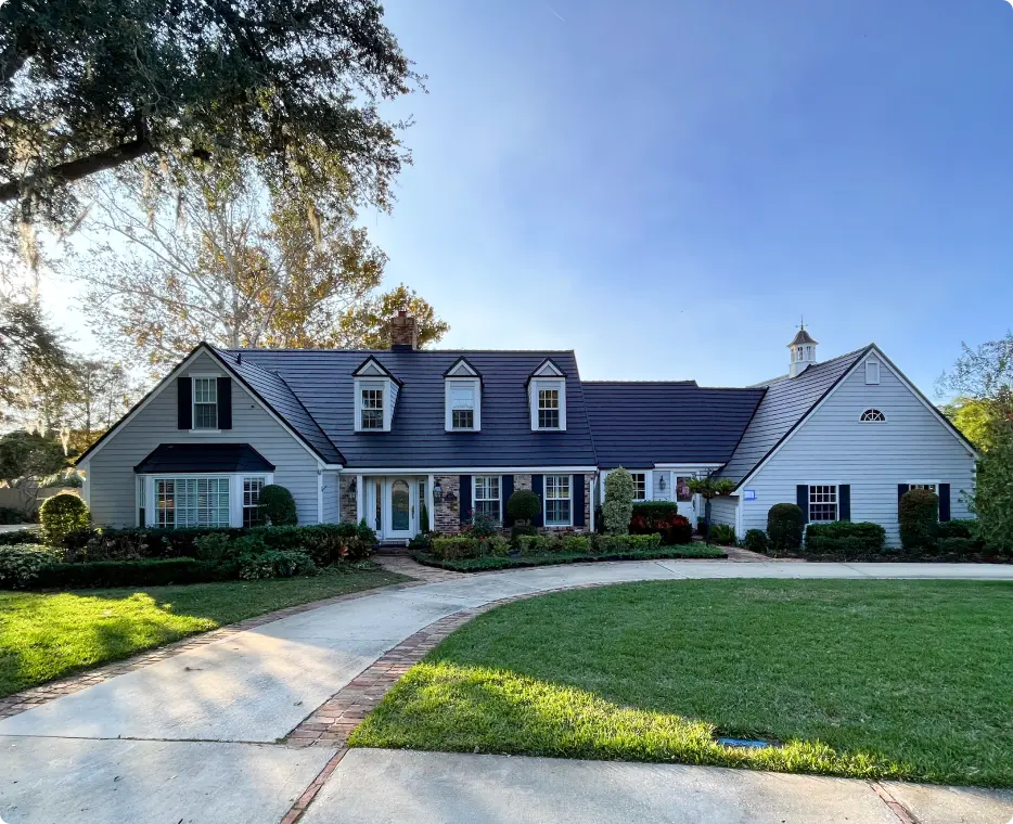 Single-story white house with dark roof, three dormer windows, manicured shrubs, and a curved driveway in front.