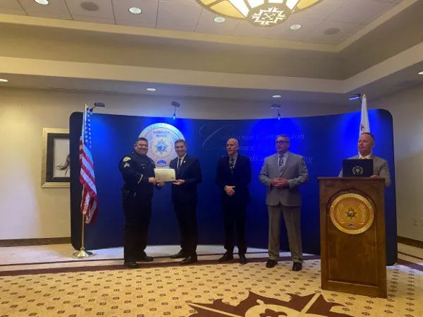 Police officer receiving a certificate from a man during an official ceremony with three other men standing on stage and American and California flags in the background.