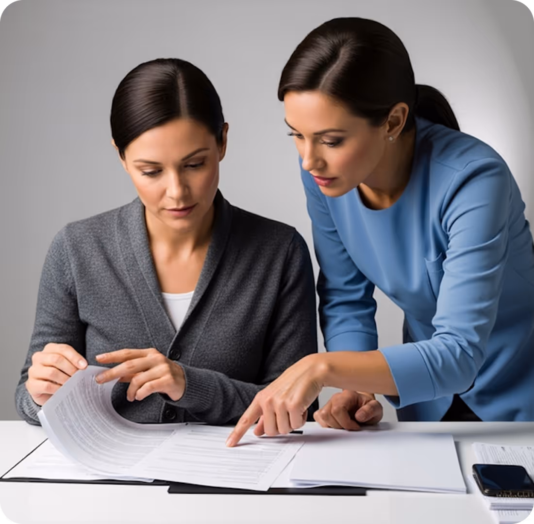 Two women reviewing and discussing documents at a white table, one pointing to the papers and the other turning pages.