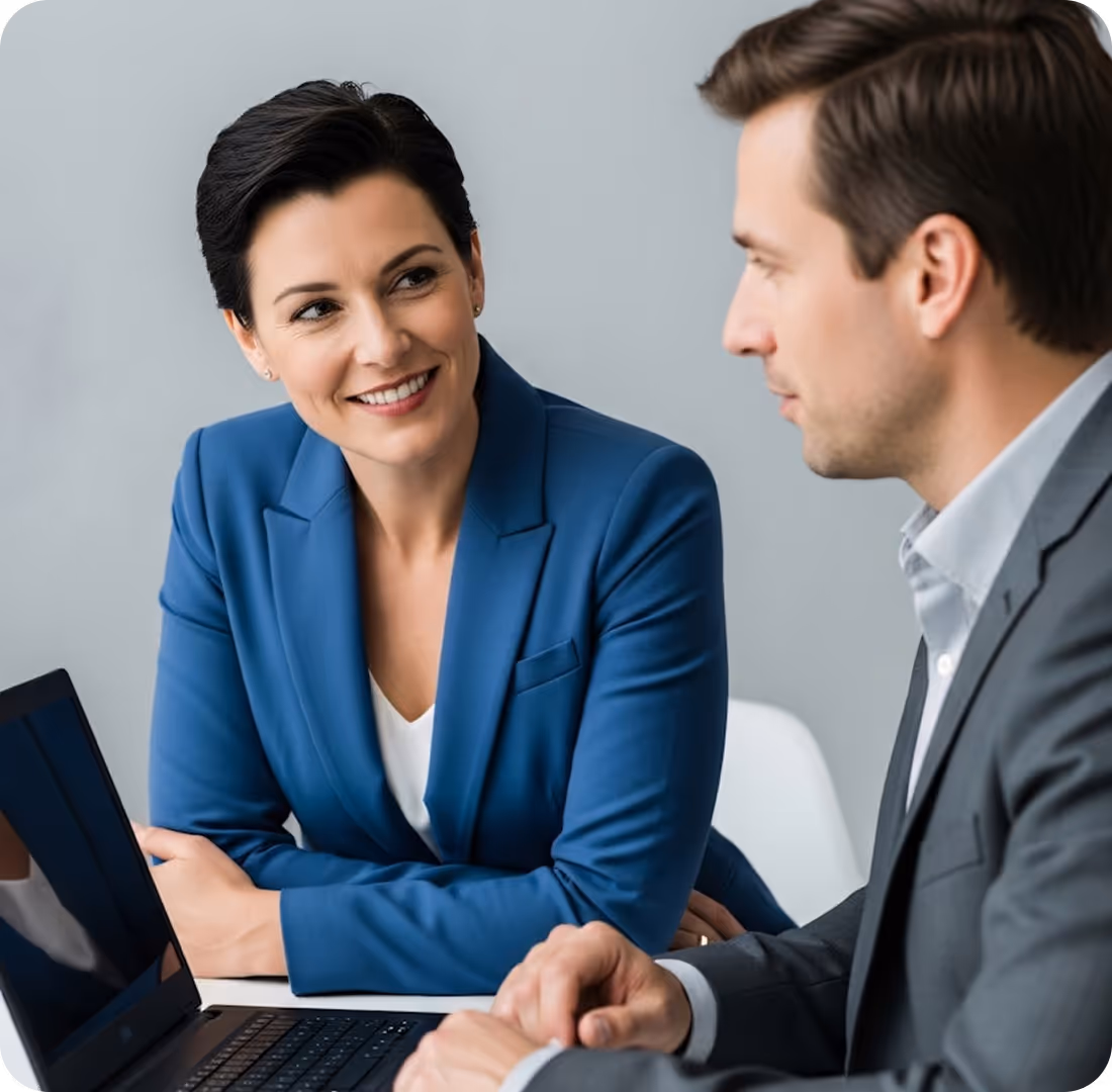 Two business professionals, a woman in a blue blazer smiling at a man in a gray suit, sitting at a table with a laptop.