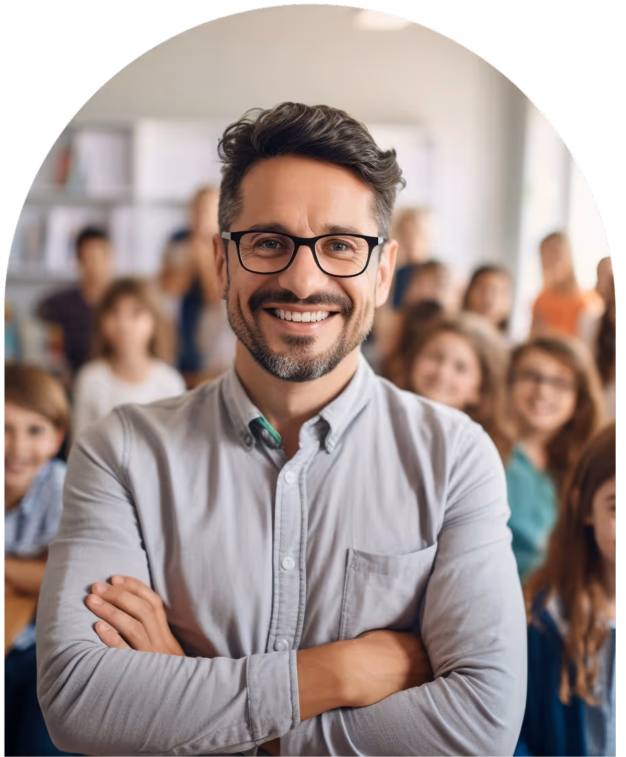 Smiling man with glasses and crossed arms standing in front of a blurred group of children in a classroom.