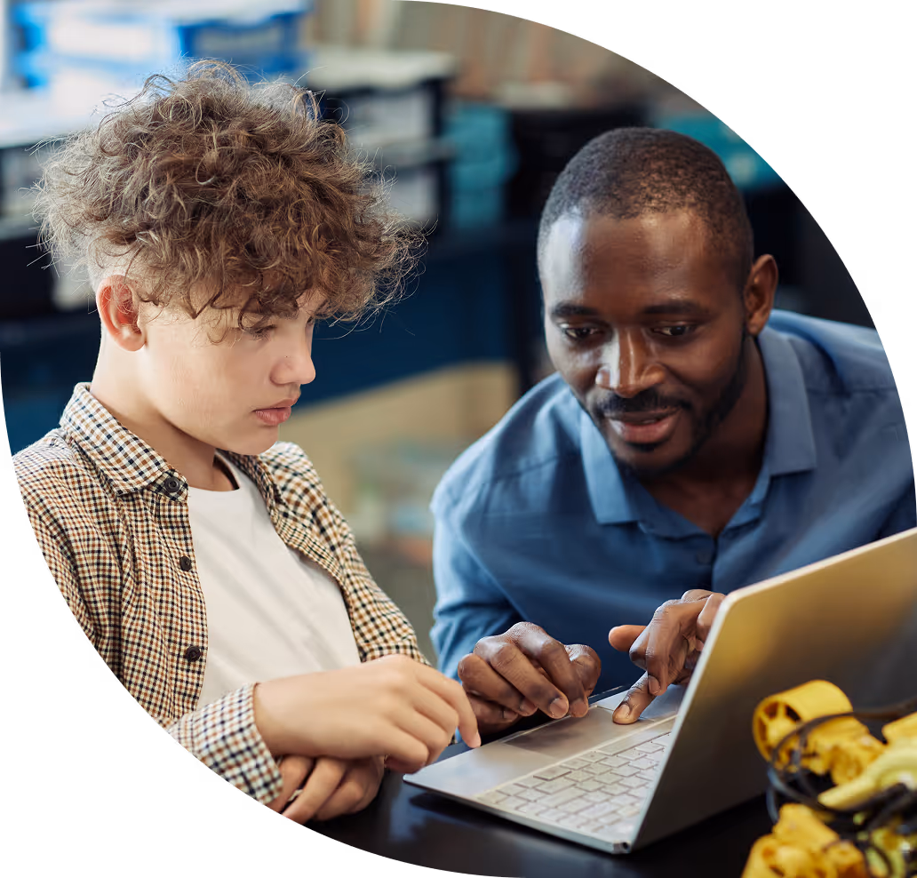 Adult man teaching a teenage boy using a laptop in a classroom setting.