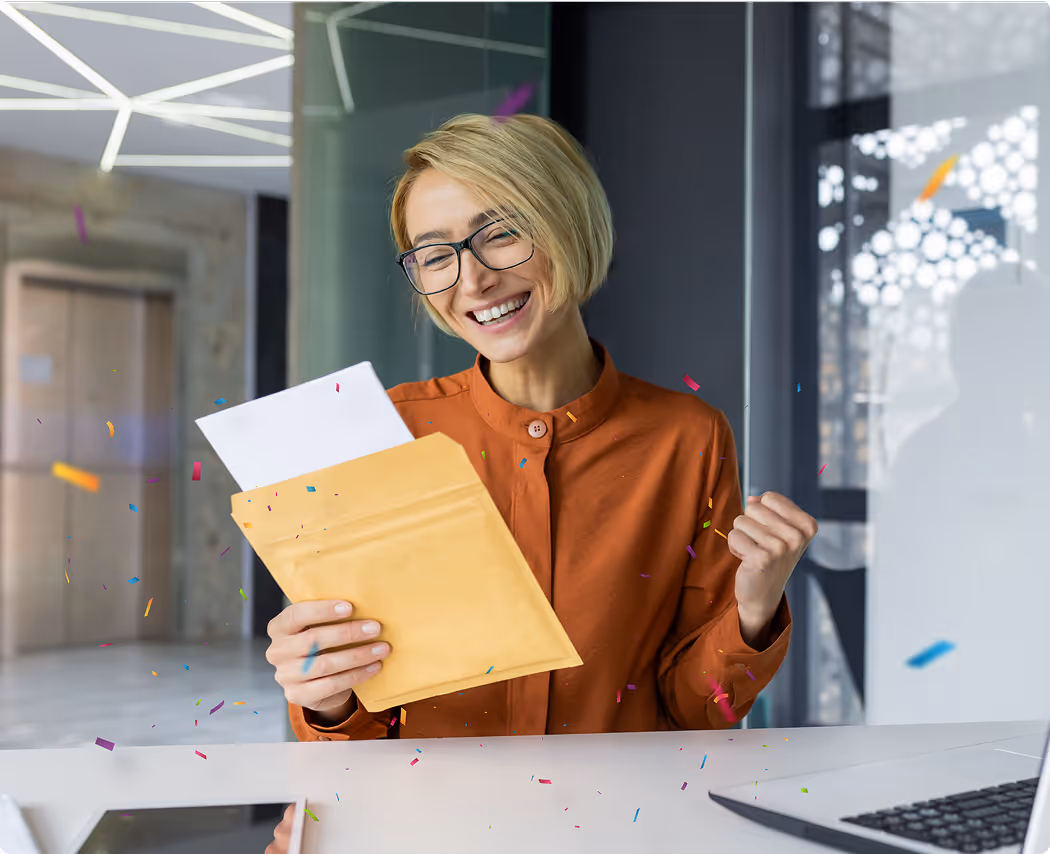 Smiling woman with short blonde hair and glasses opening a yellow envelope and celebrating at a desk with confetti around her.