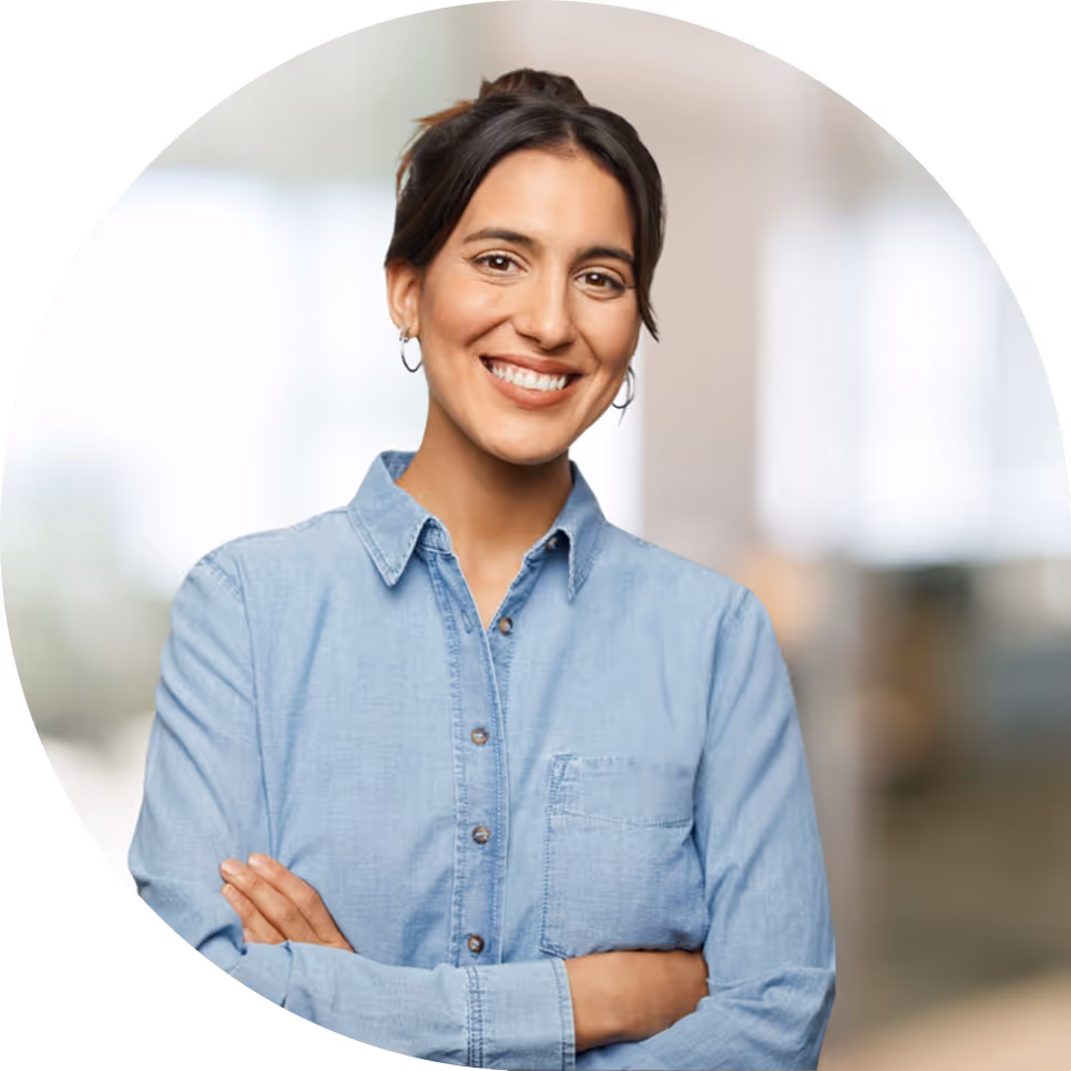 Smiling woman with dark hair wearing a light blue denim shirt standing with arms crossed in a bright indoor setting.