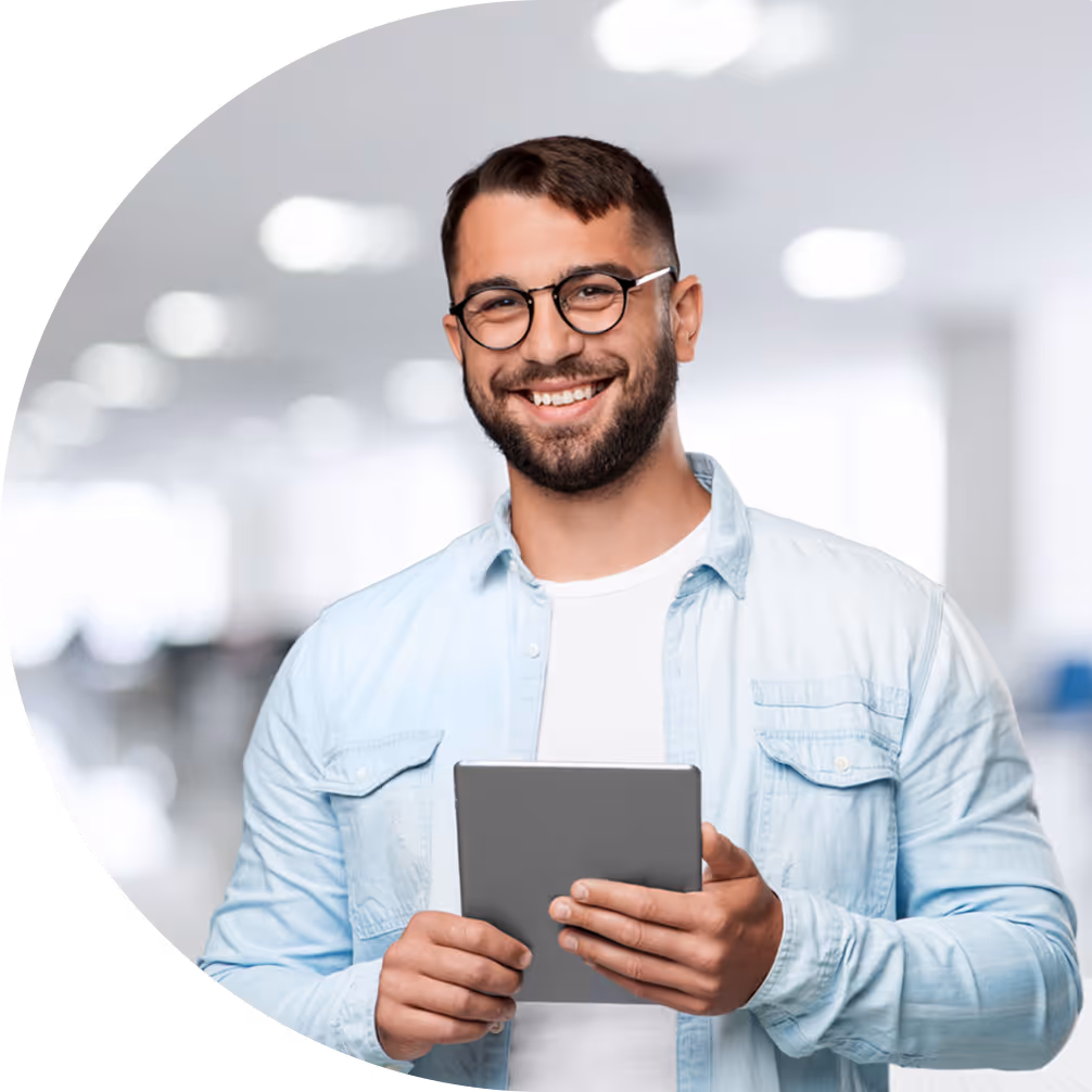 Smiling man wearing glasses and light denim shirt holding a tablet in a bright office.