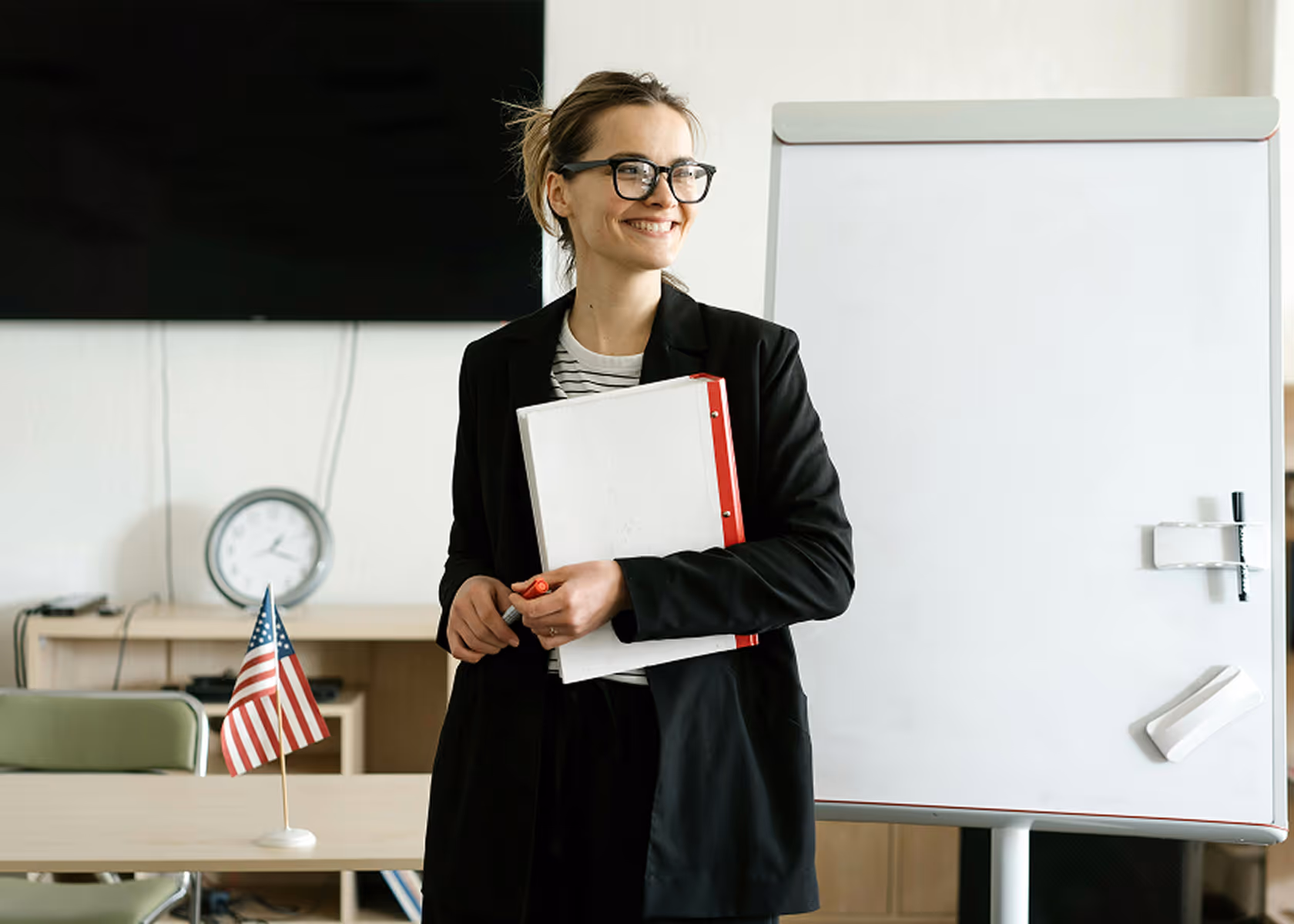 Smiling woman in glasses holding a white binder and marker standing by a blank flip chart in an office with a small American flag on a desk.