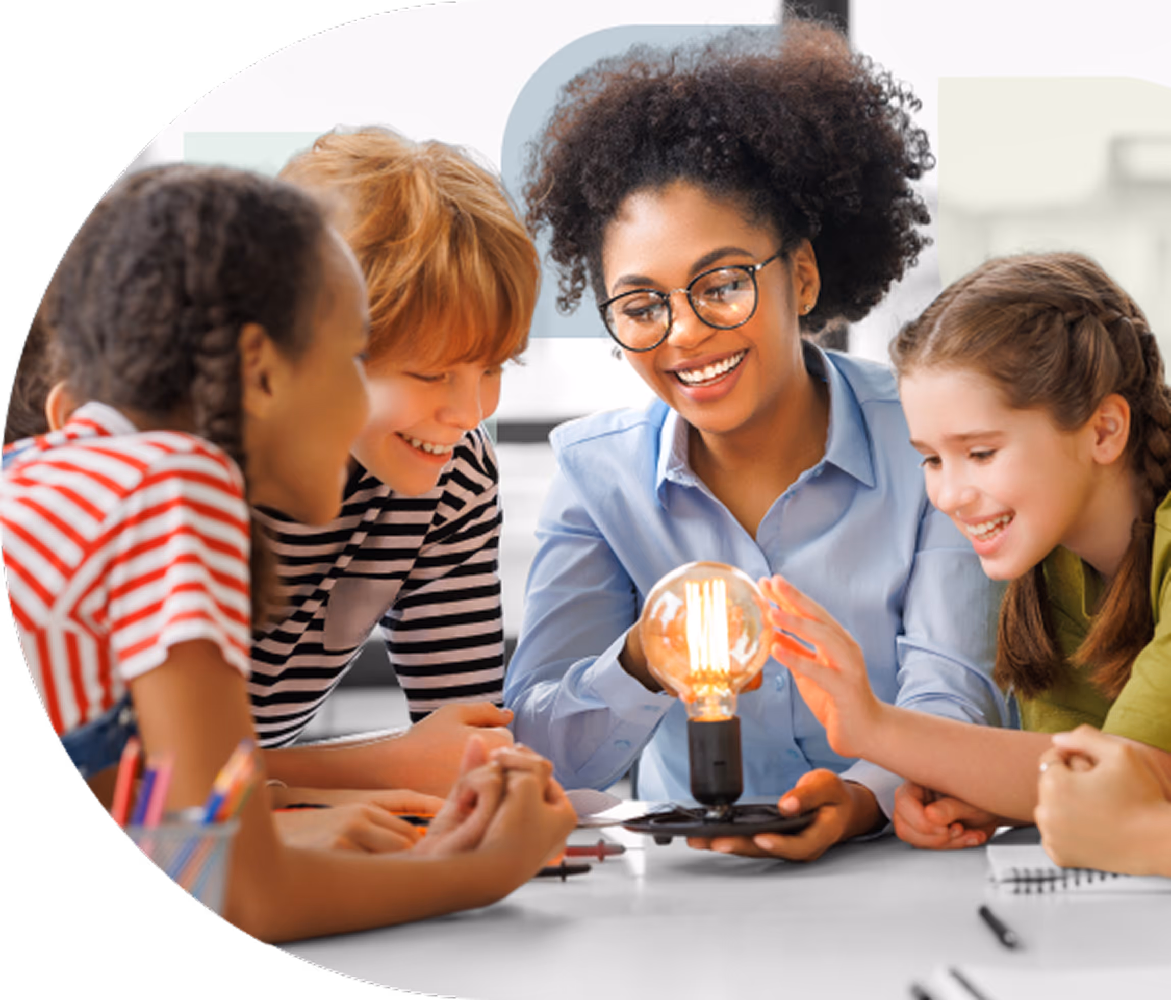 A teacher with curly hair and glasses shows a glowing light bulb to a group of smiling children gathered around a table.
