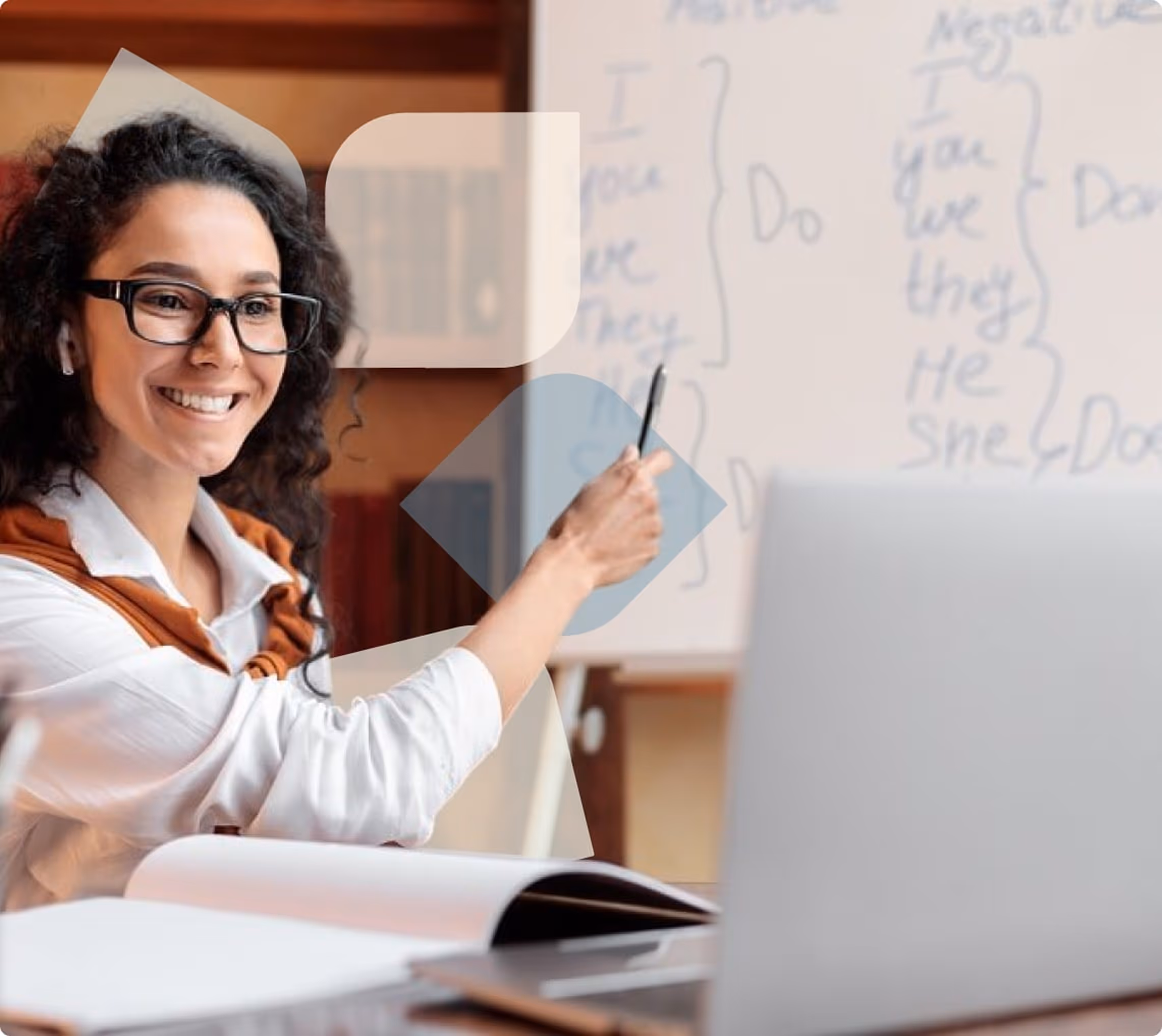 Smiling woman with glasses pointing to a whiteboard while sitting at a desk with an open book and laptop during an online lesson.