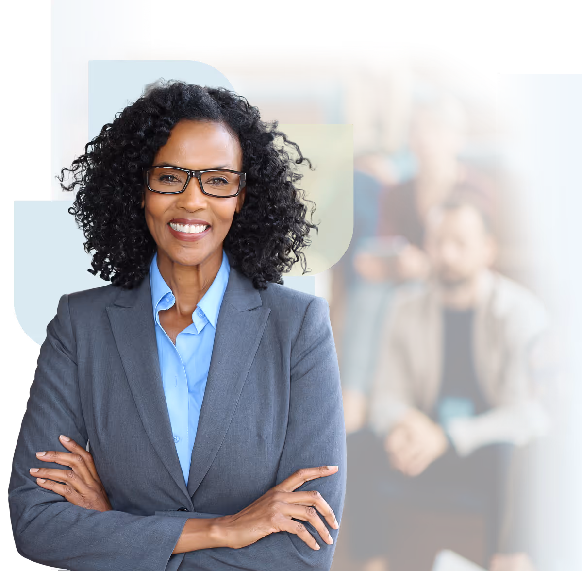 Confident woman with curly hair and glasses smiling with her arms crossed, with blurred people in the background.