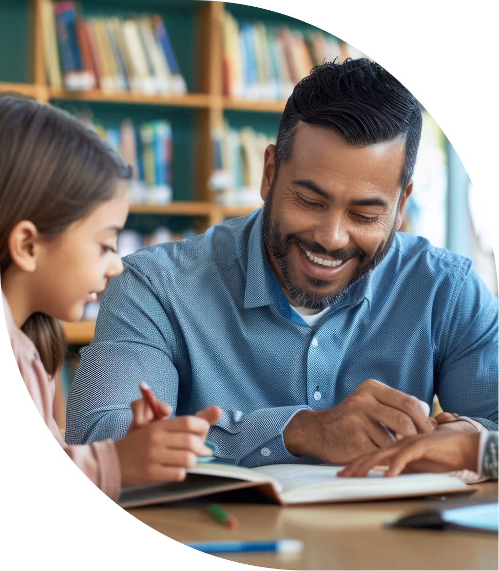 Smiling man helping young girl with reading in a library or classroom setting.